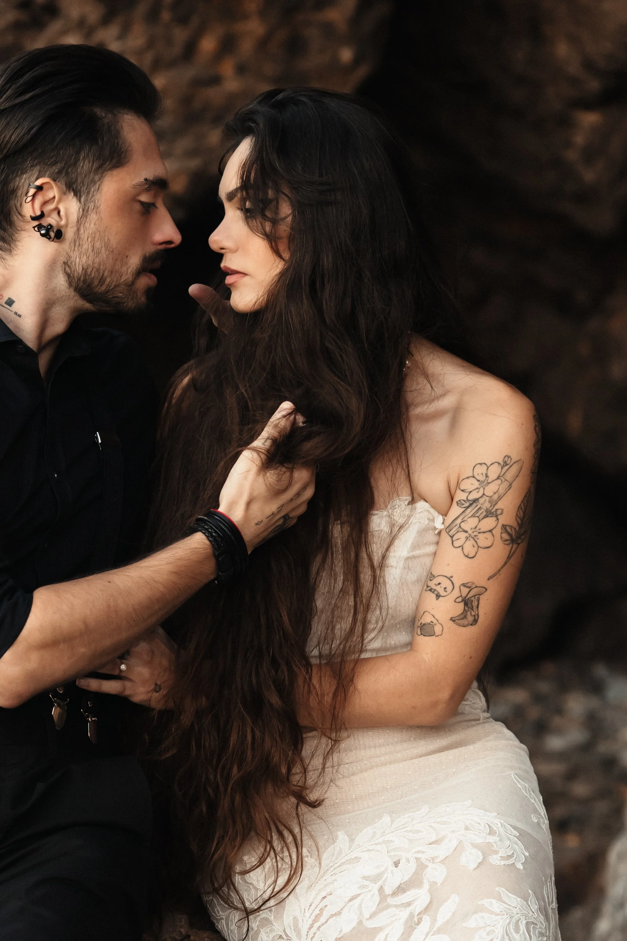 Un homme et une femme se regardent intensément, près d'une paroi rocheuse, l'homme a plusieurs piercings et une barbe, la femme a de longs cheveux foncés et des tatouages sur le bras et l'épaule, elle porte une robe blanche avec des motifs floraux.