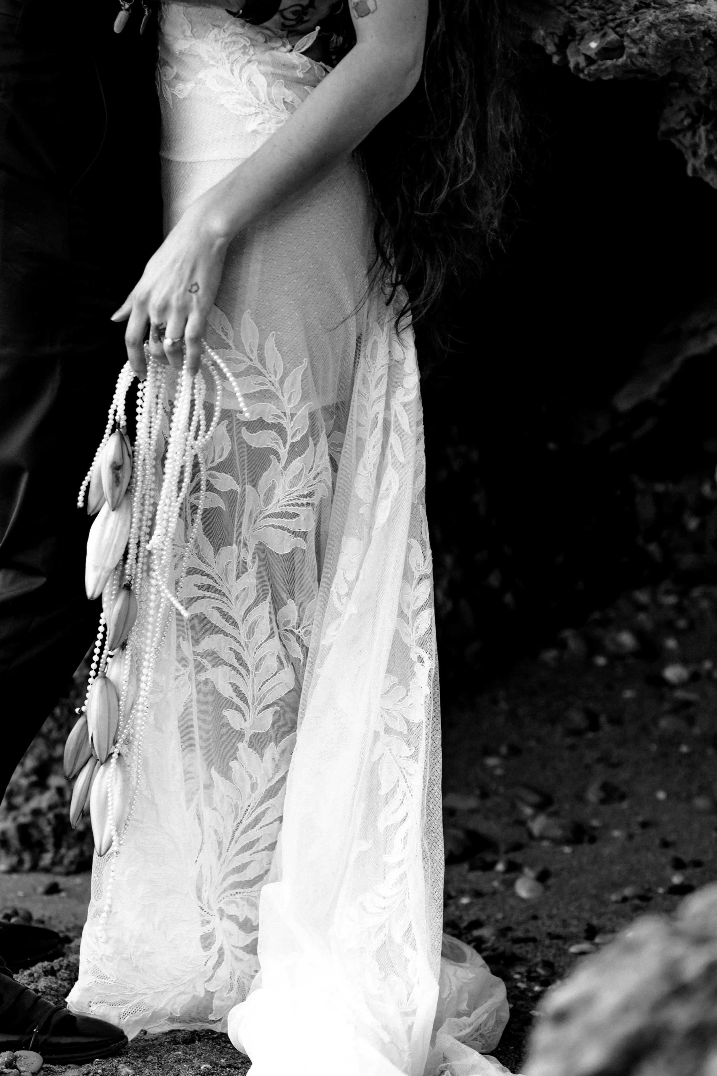 Détail d'une robe de mariée en dentelle avec des feuilles brodées, une main tenant des colliers de perles et de coquillages, dans un environnement extérieur.
