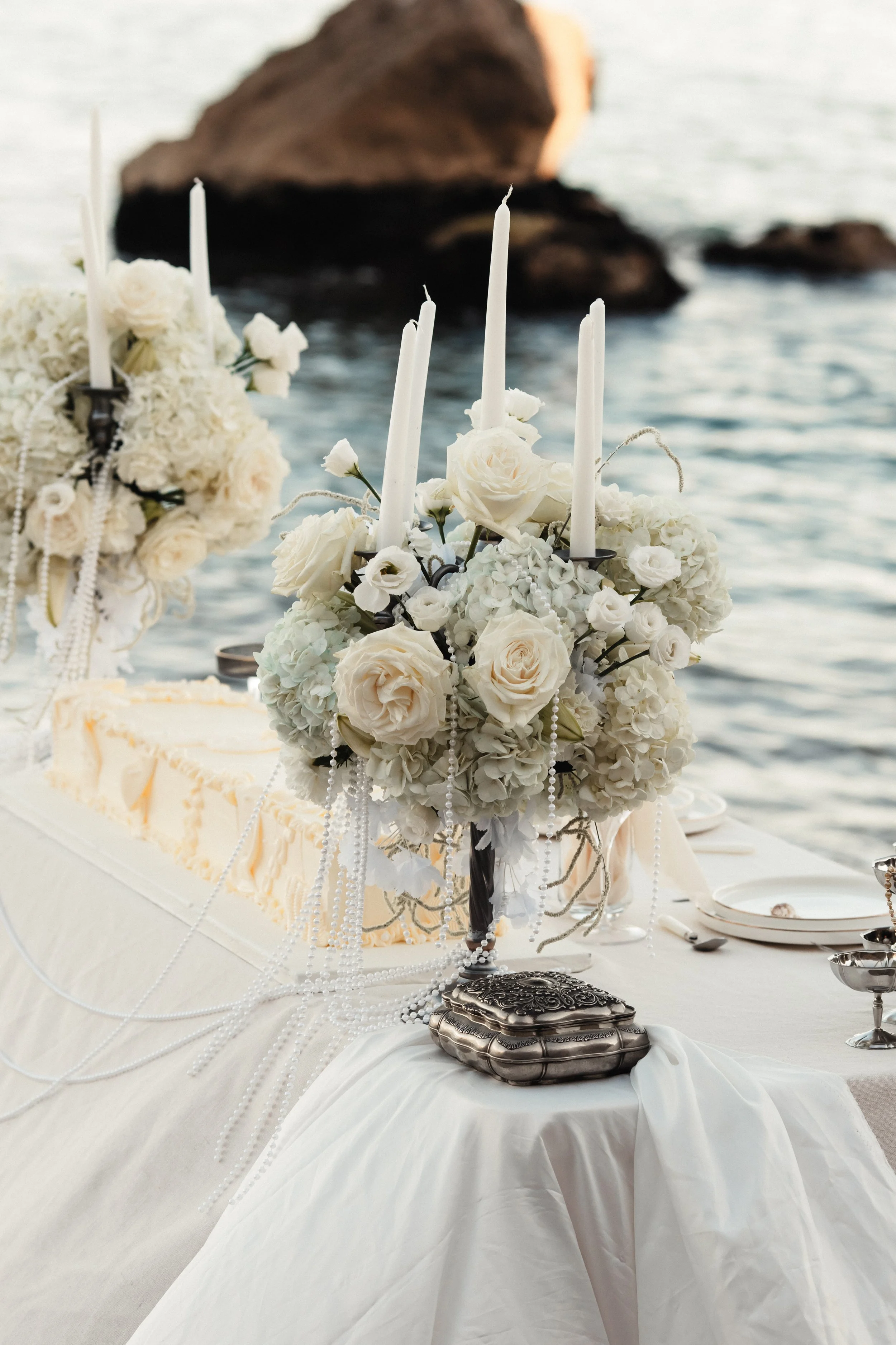 Table décorée avec des bougies, des fleurs blanches, et des perles, près de la mer.