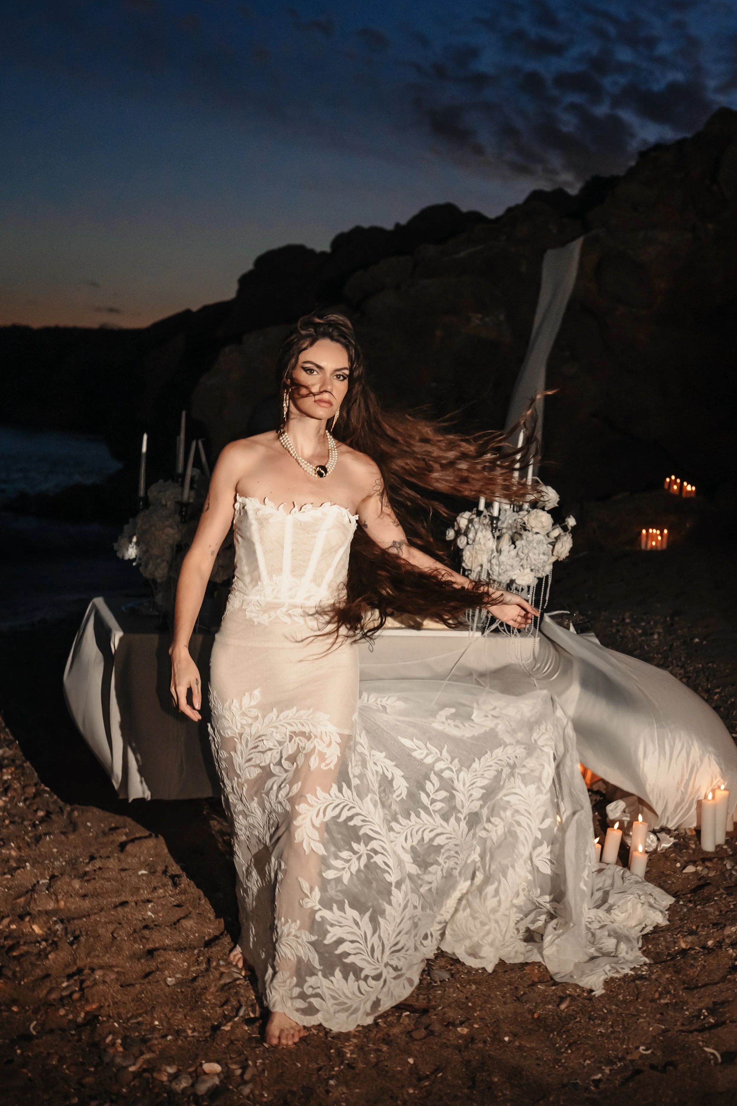 Femme en robe de mariée sur une plage au crépuscule, projection romantique avec bougies et décor floral.