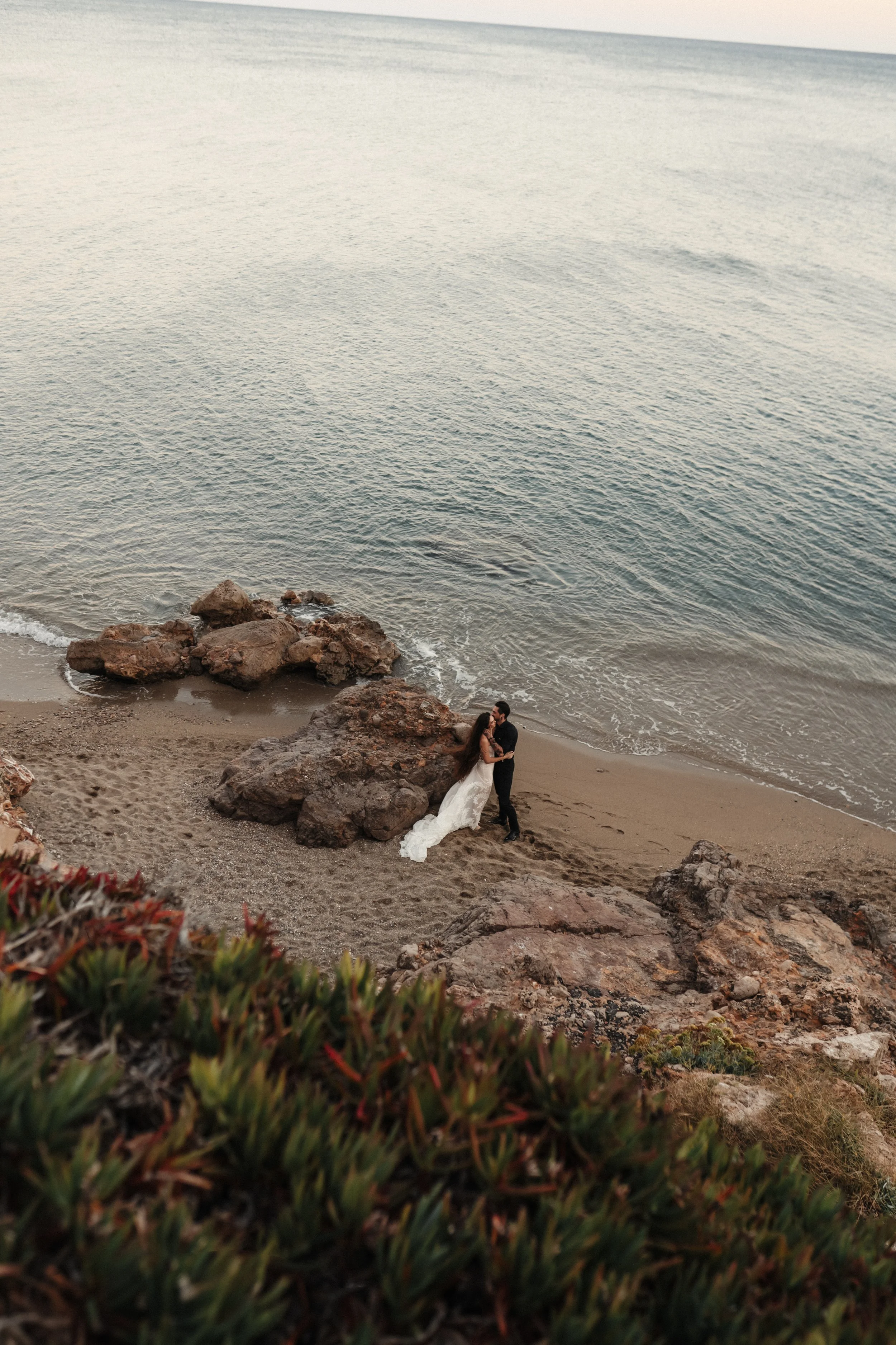 Un couple en mariage sur la plage, entouré de rochers, avec la mer en arrière-plan.