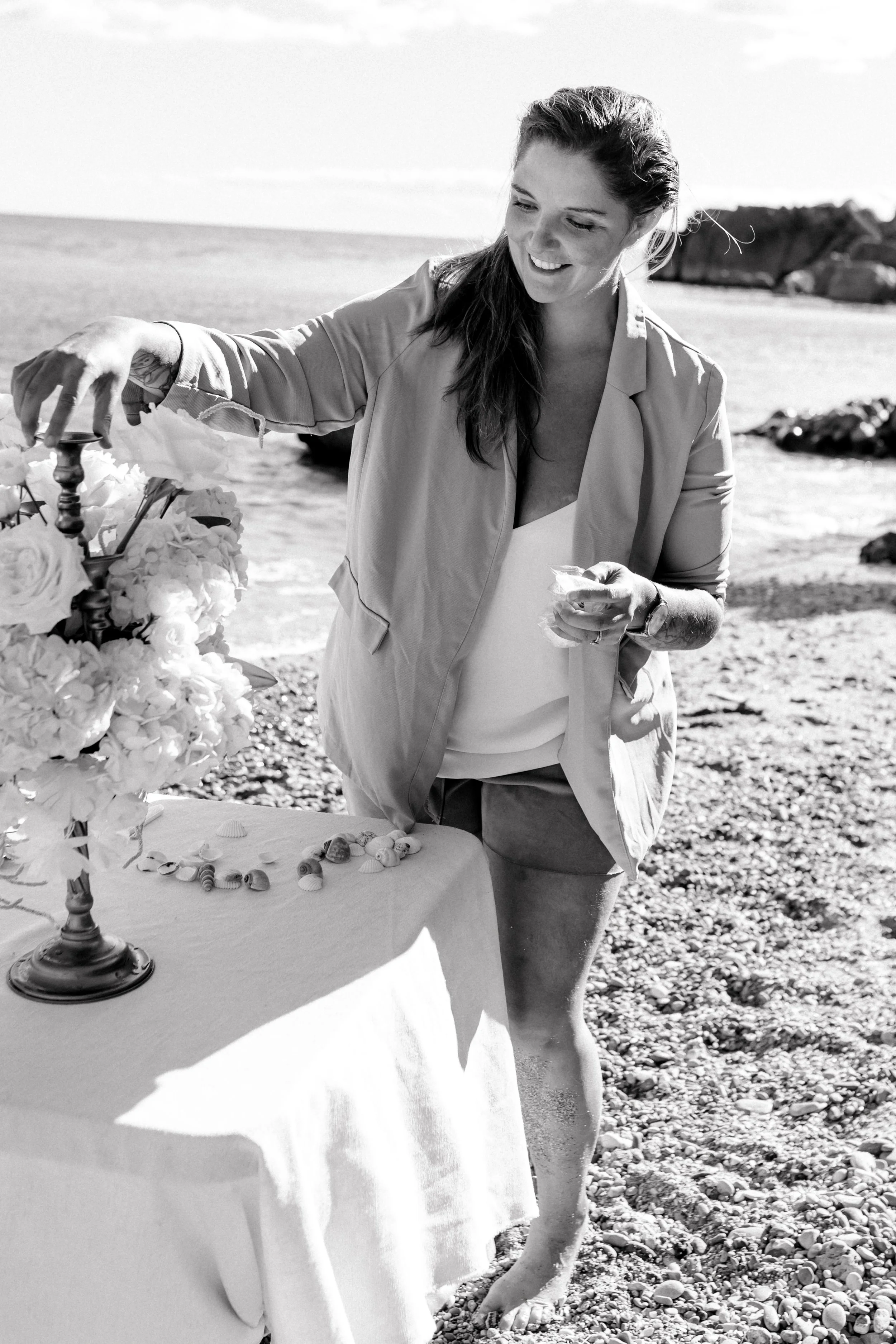 Une femme souriante décore une table avec des fleurs et des coquillages sur une plage, en noir et blanc.