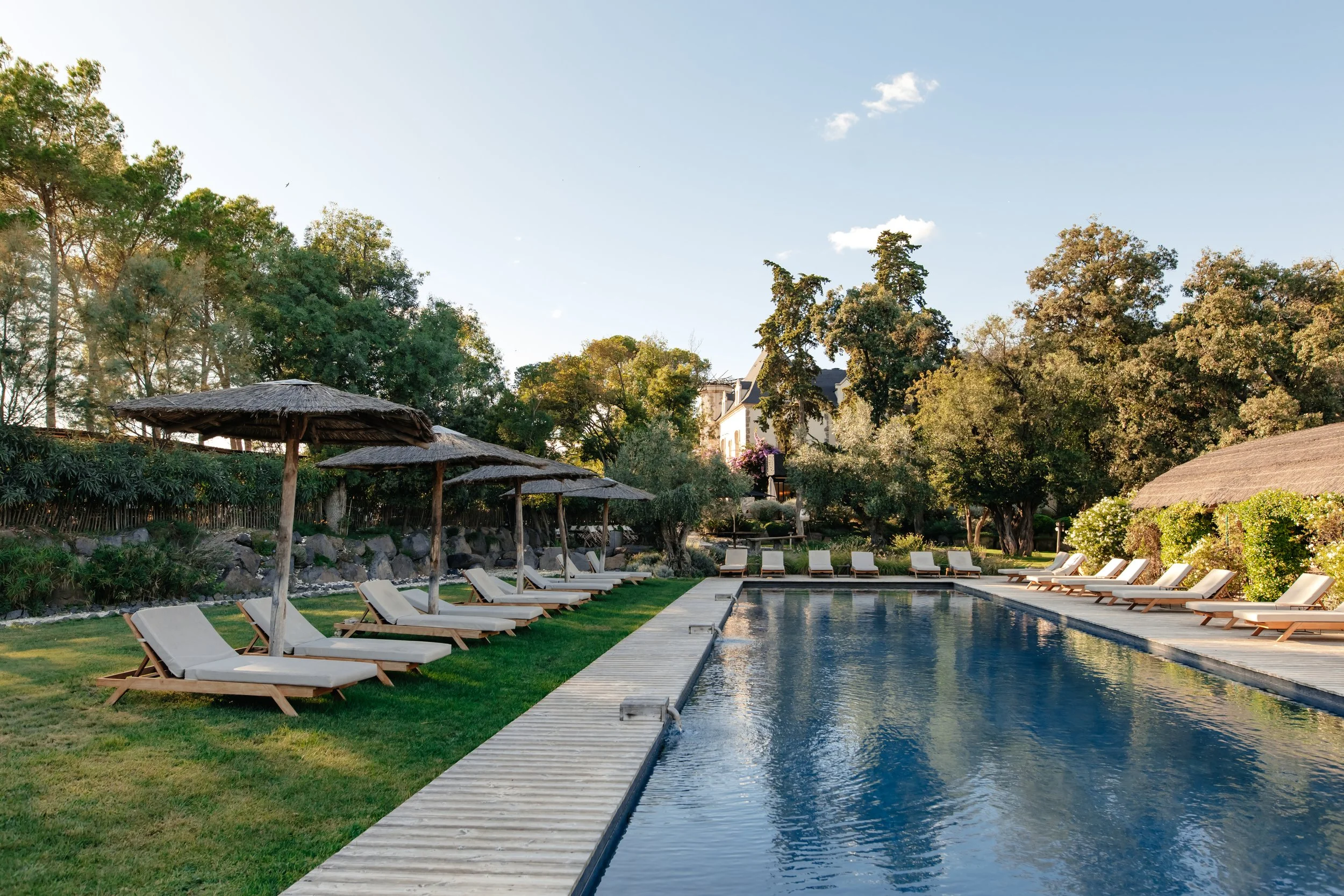 Piscine extérieure avec des transats en bois et des parasols en paille, entourée de verdure et d'arbres, sous un ciel clair.