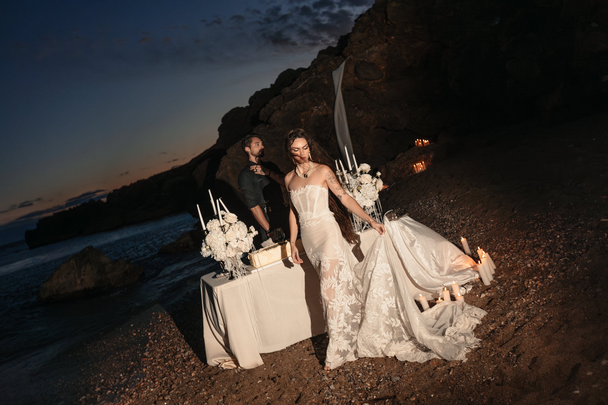 Un couple en tenue de mariage sur une plage rocheuse au coucher du soleil, avec des bougies et des décorations blanches, une femme en robe de mariée white avec longues cheveux bruns, un homme en vêtement noir, ambiance romantique et artistique.
