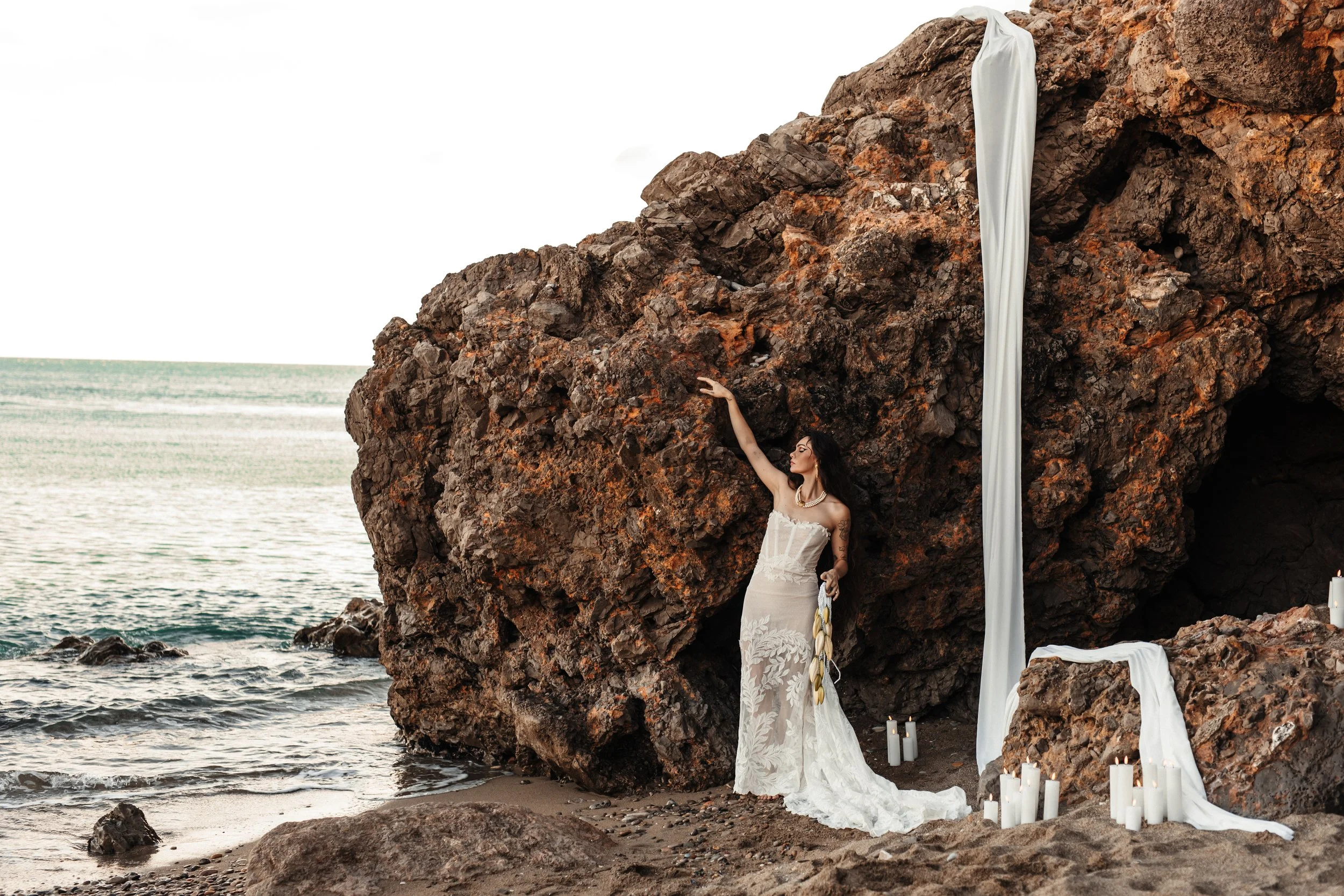Une femme en robe de mariée se tenant près d'un rocher sur la plage, avec des bougies autour et un drapé blanc suspendu au rocher.