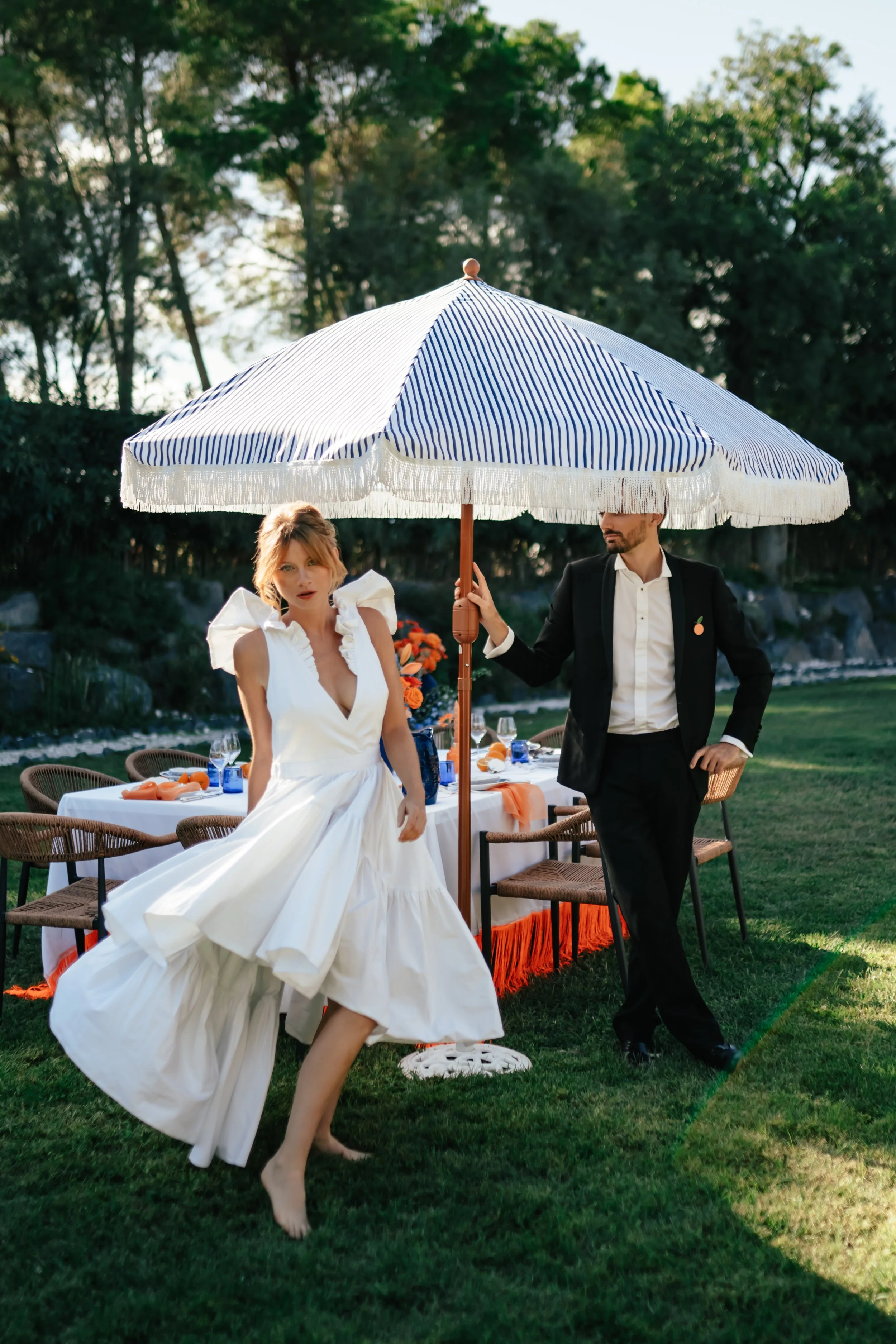 Une femme en robe blanche à la jupe volumineuse, et un homme en costume noir, se tiennent sous un grand parasol rayé blanc et bleu, près d'une table de fête décorée avec des verres et des assiettes, en plein air, avec des arbres en arrière-plan.