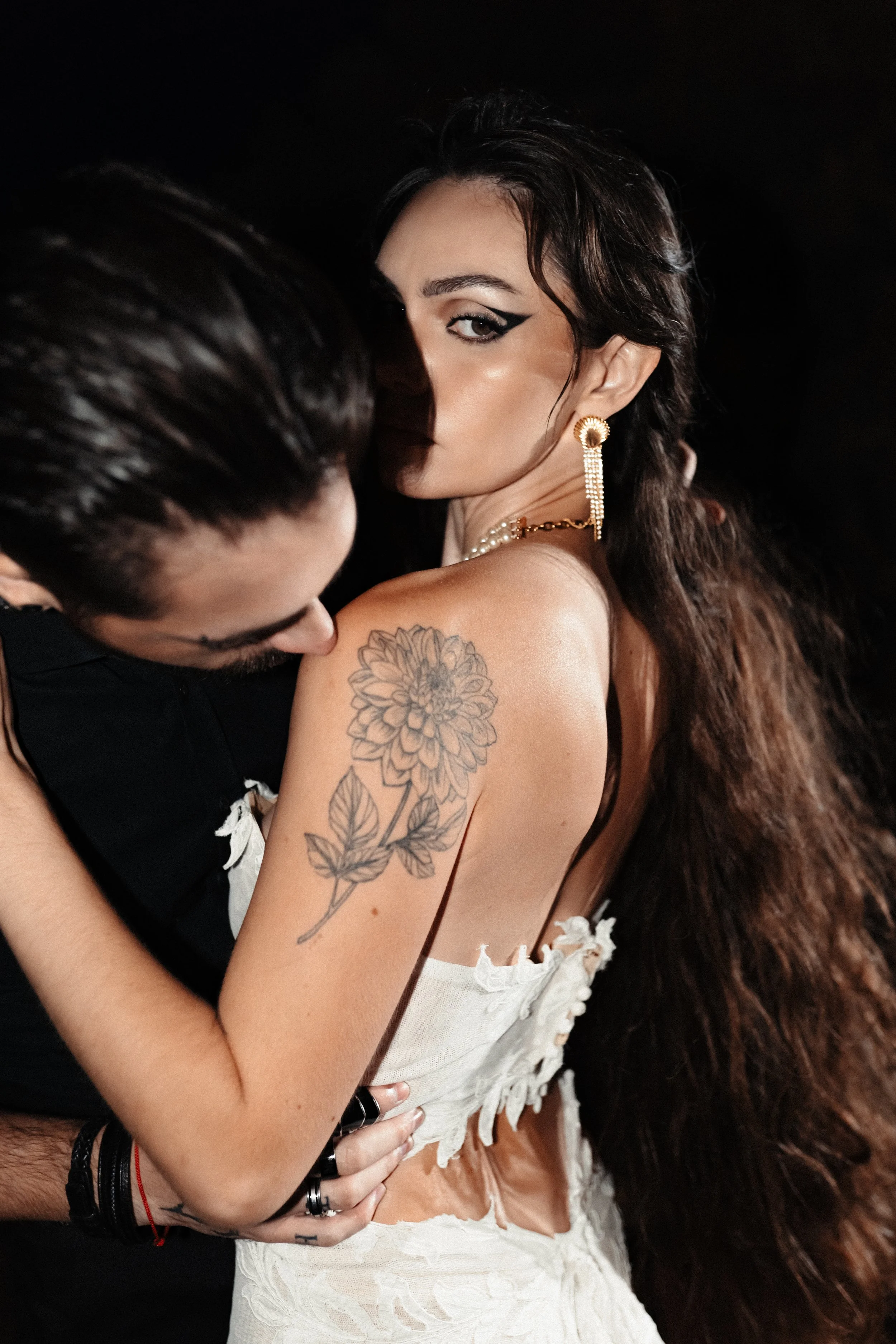 Une femme avec une grosse tatouage de fleur sur l'épaule, portant des bijoux dorés, se fait embrasser par un homme dont le visage est en partie visible, contre un fond sombre.