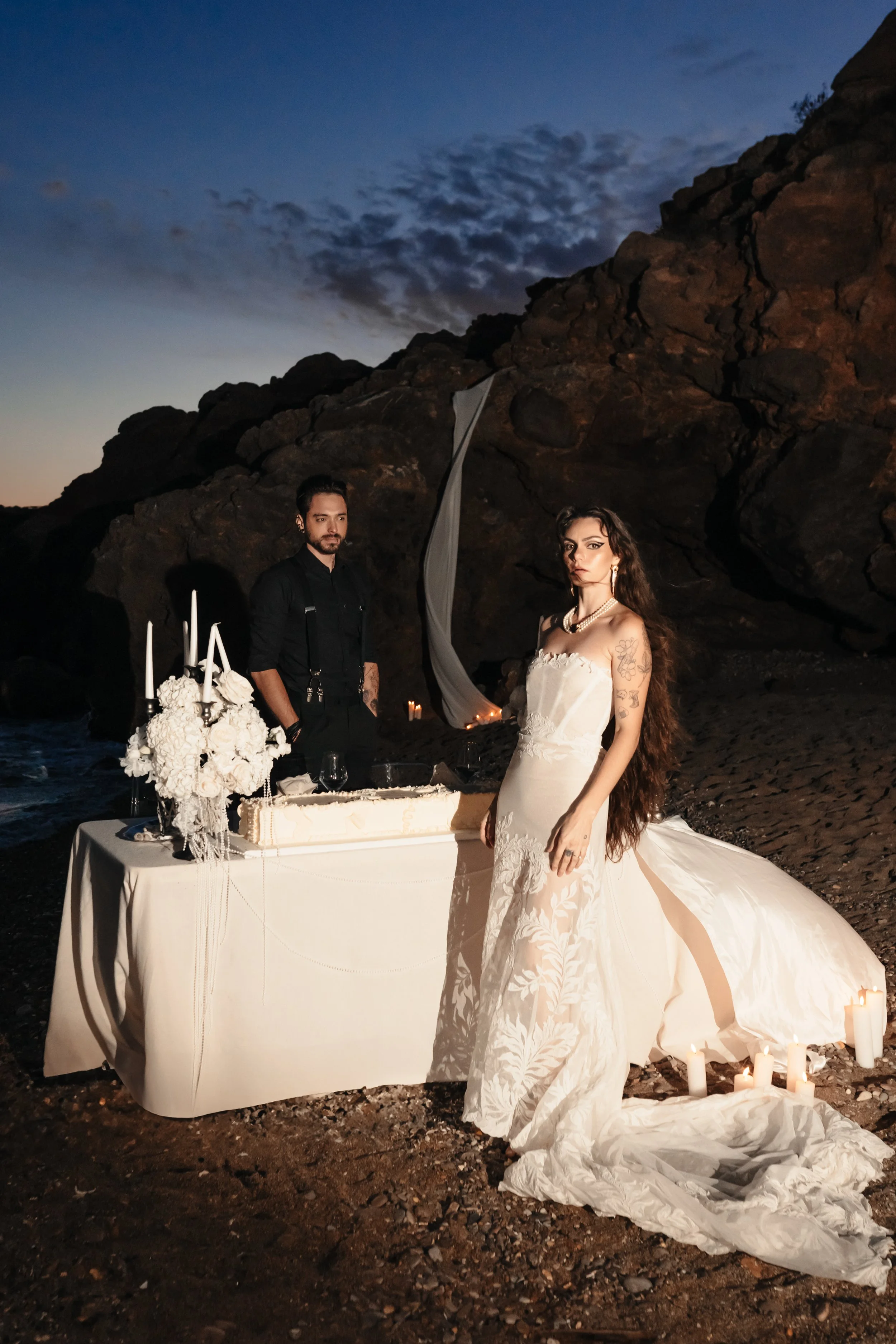 Mariée en robes blanche et garçon d'honneur ou invité se tenant près d'une table de mariage sur une plage rocheuse au crépuscule.