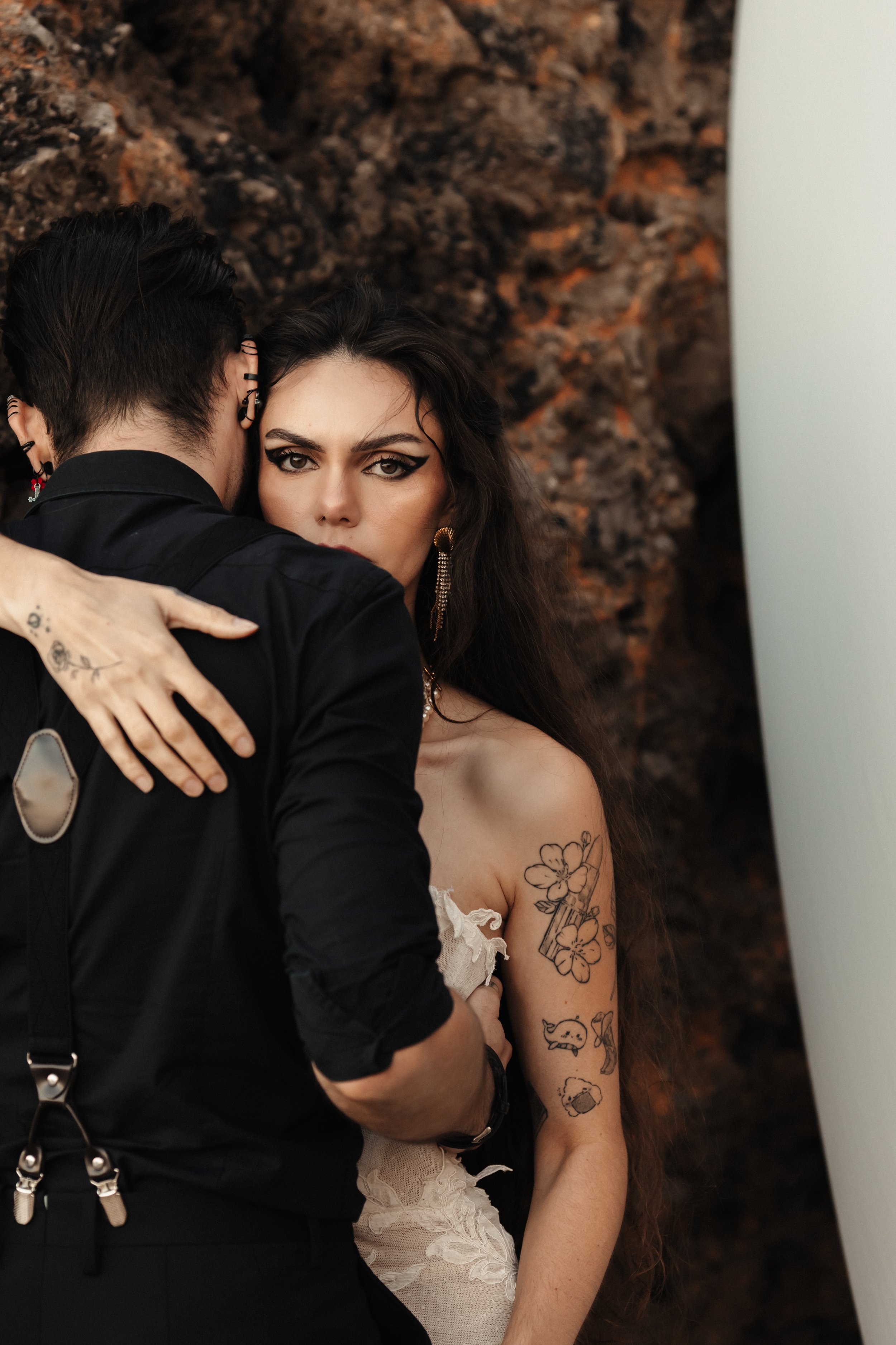 Un couple en pose artistique, l'homme ayant des cheveux noirs et portant une chemise noire, et la femme avec de longs cheveux bruns et maquillage noir prononcé, la femme porte une robe beige ornée de tatouages visibles sur son bras, dans un décor roc