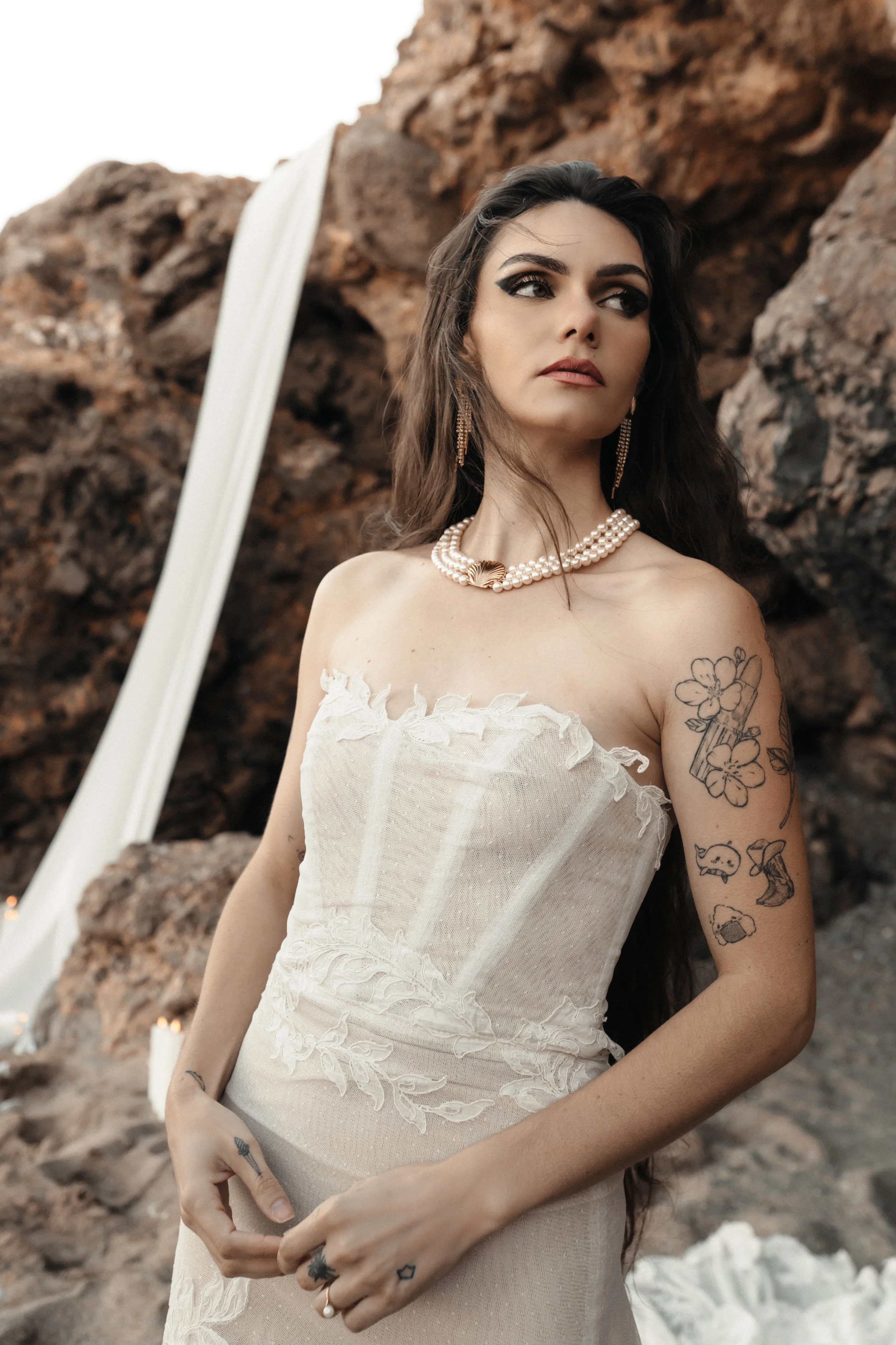 Jeune femme avec des tatouages portant une robe blanche en tissu léger, des bijoux en perles, et des boucles d'oreilles, posant devant un fond rocheux et un couloir d'eau en forme de cascade.