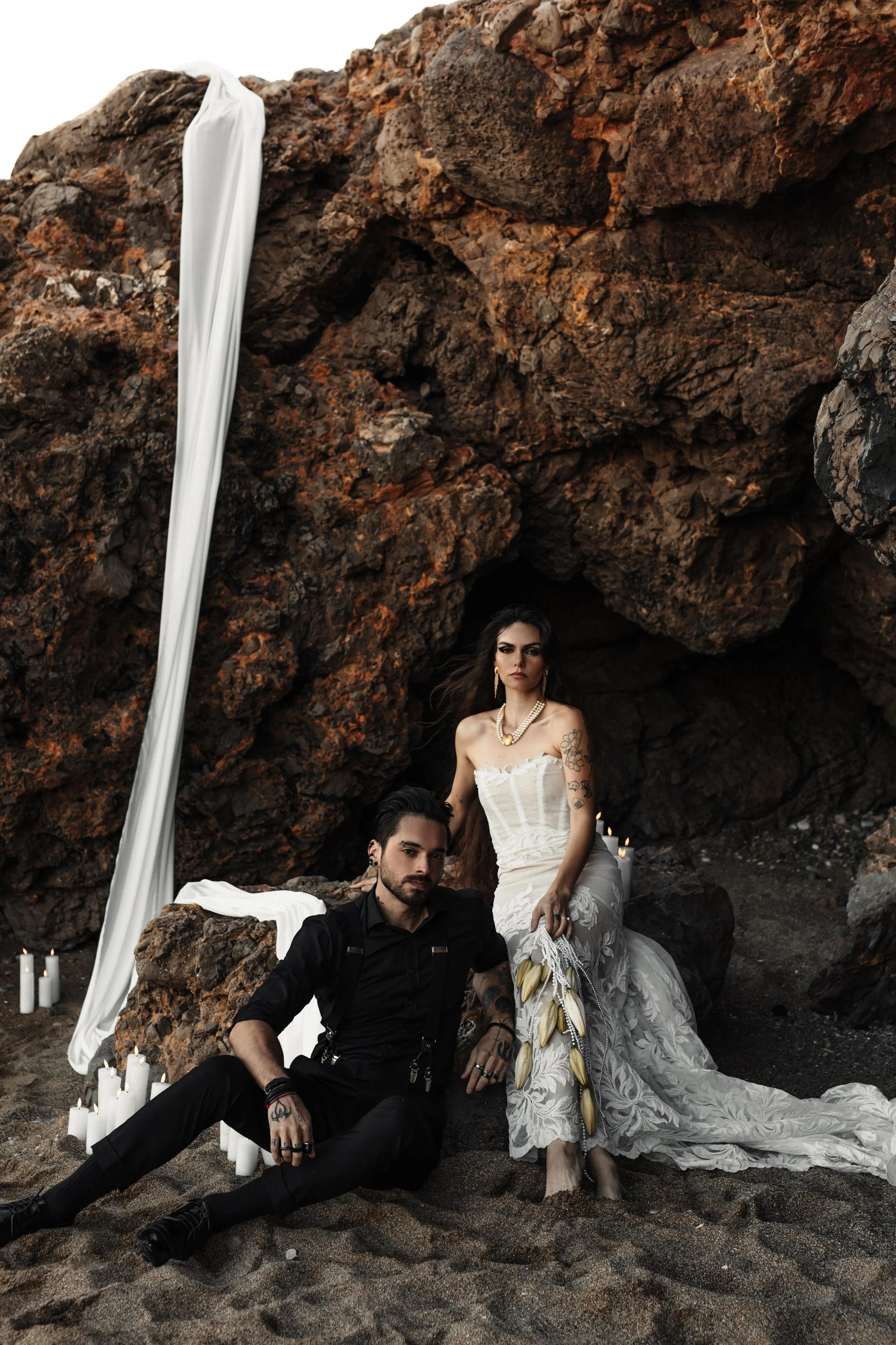 Un couple posant sur une plage avec une roche en arrière-plan. La femme porte une robe blanche de style vintage et des boucles d'oreilles dorées, et le homme est habillé en noir avec des tatouages visibles, des accessoires et une barbe, entourés de b