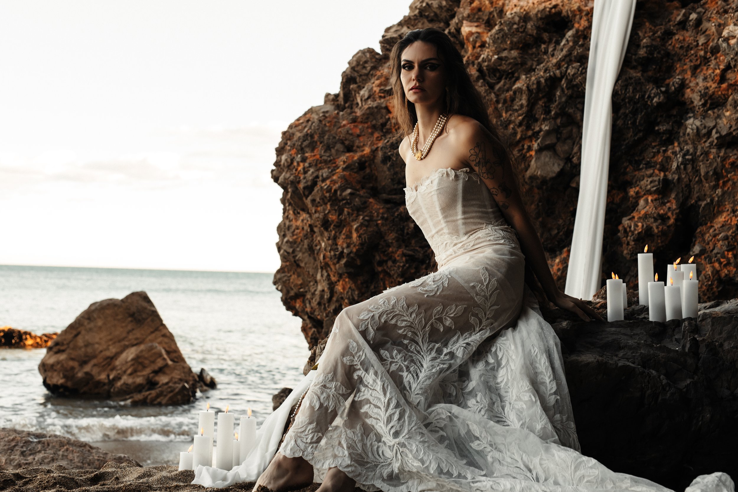 Femme en robe blanche élégante assise sur des rochers au bord de la mer, entourée de bougies allumées, avec un décor de drapé blanc et une falaise rocheuse, scène romantique au coucher du soleil.