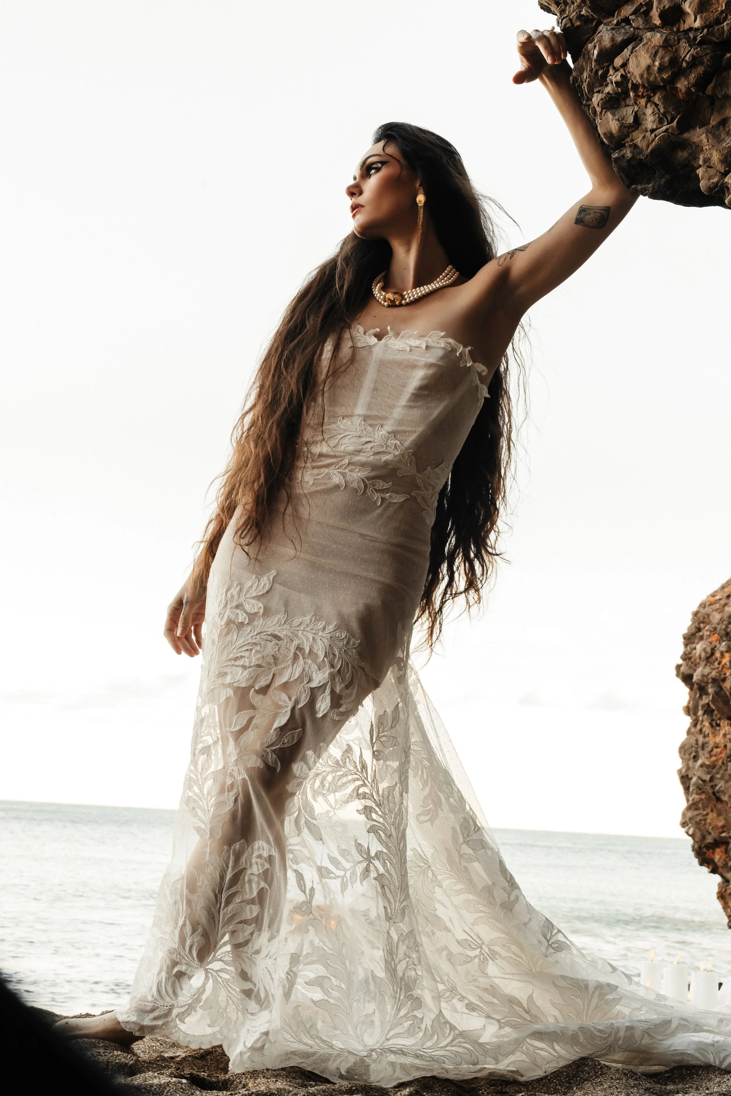 Une femme portant une robe longue en dentelle beige, avec des bijoux dorés, debout près de la mer, appuyée contre une roche.