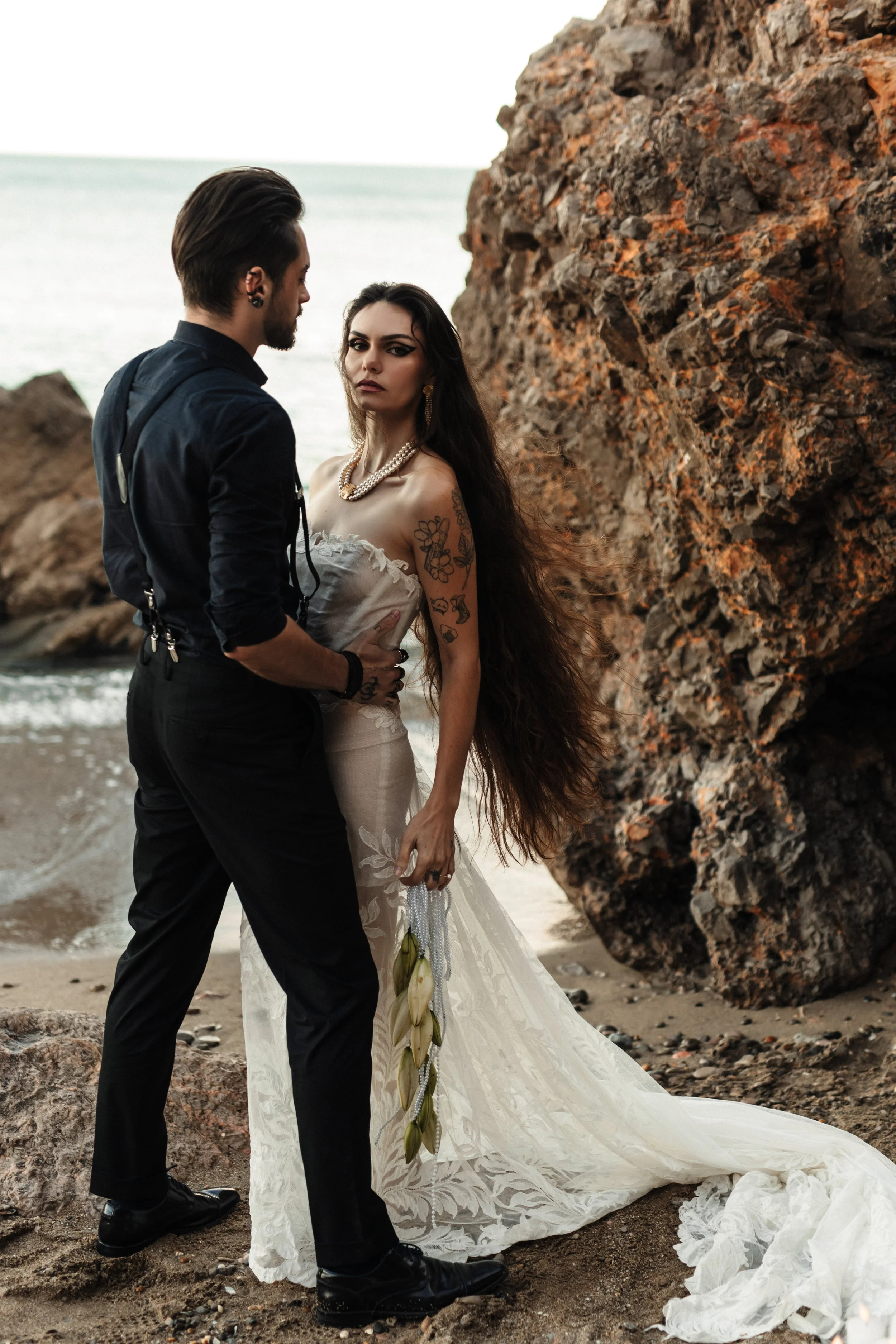 Un couple en tenue de mariage se tient près de la plage, avec grandes roches en arrière-plan, la femme porte une robe blanche avec tatouages et bijoux, le homme porte une chemise noire