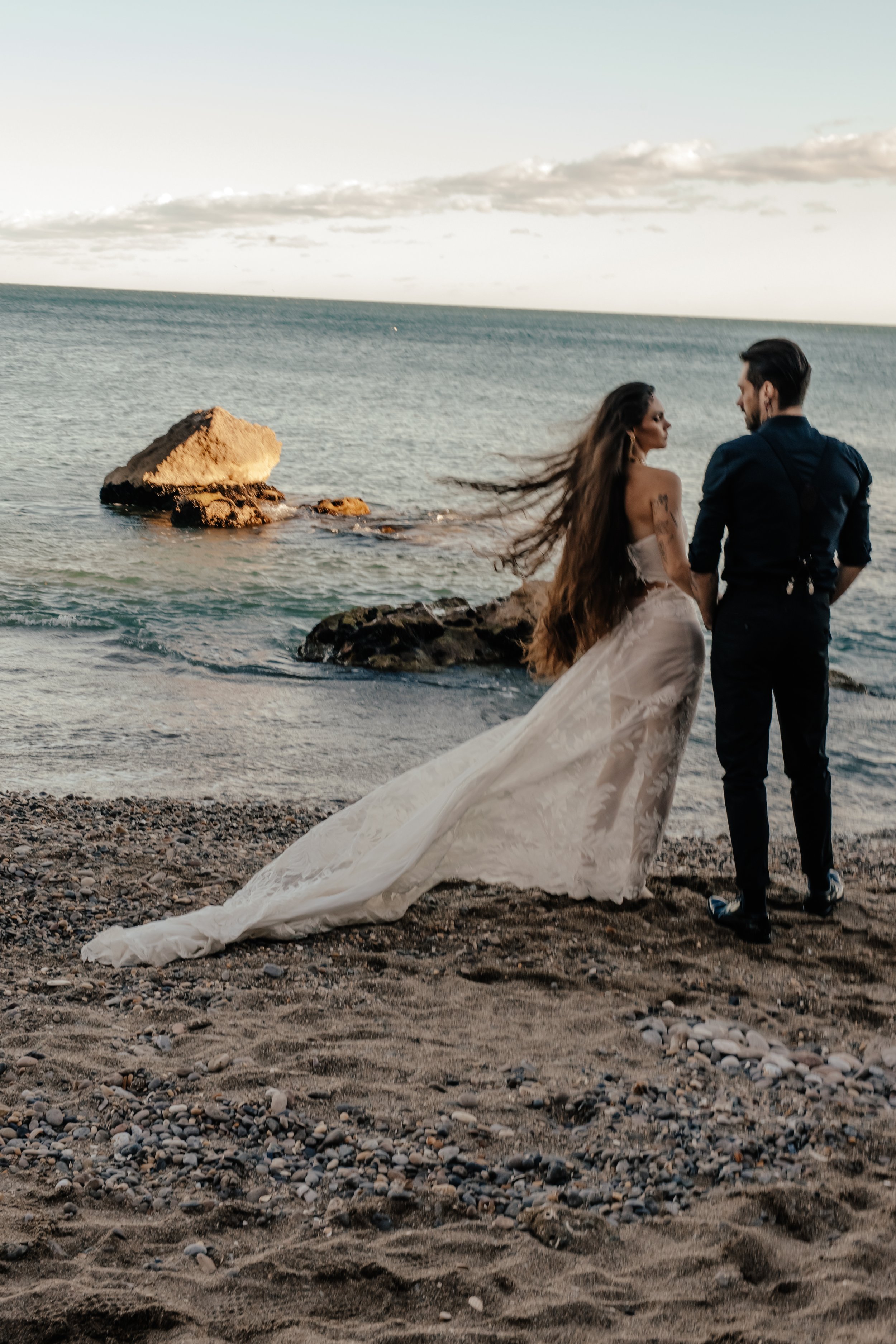 Un couple en vêtements de mariage se tenant par la main sur une plage près de la mer, avec des rochers en arrière-plan, au coucher du soleil.