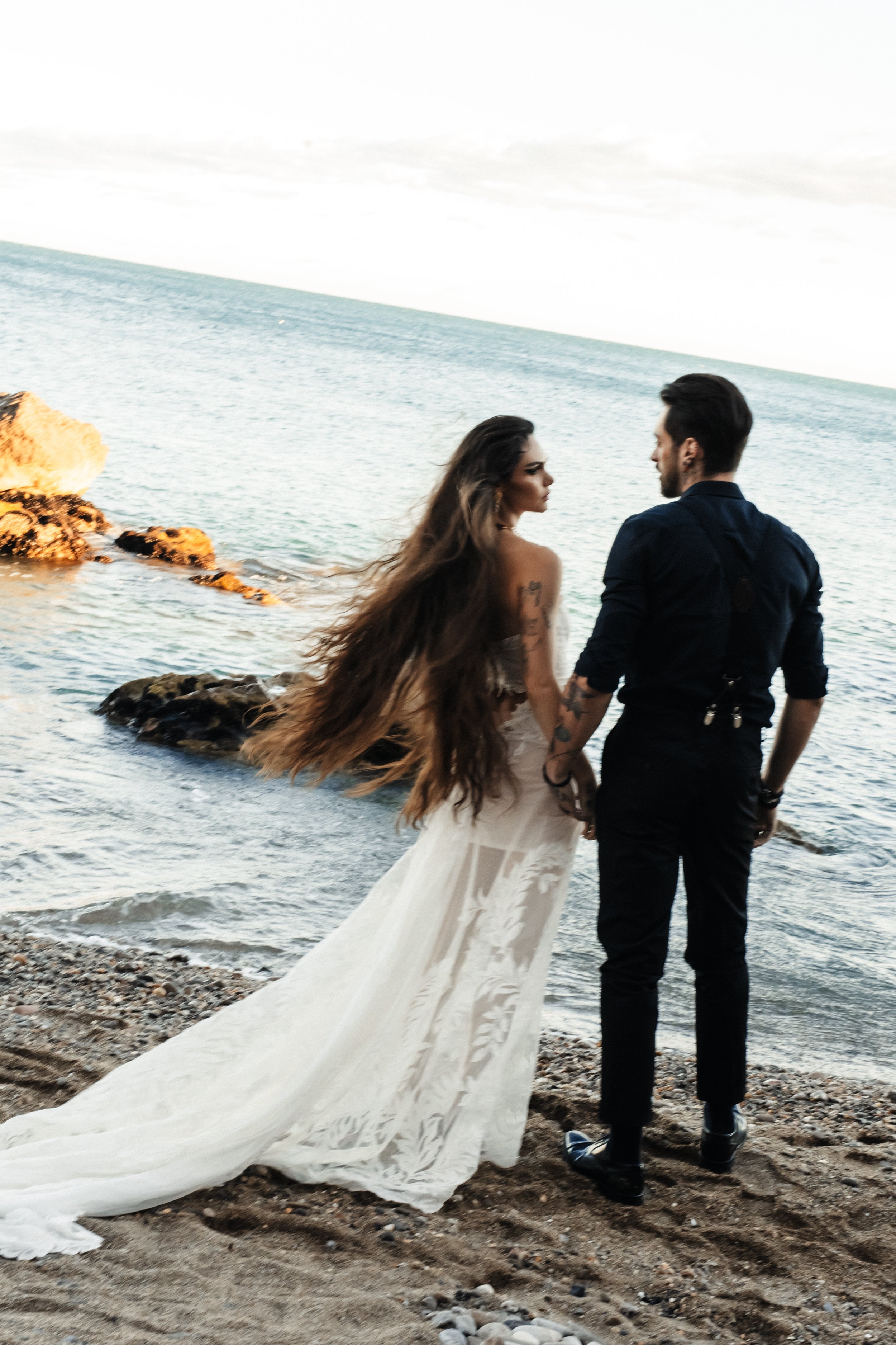 Un couple debout sur la plage près de la mer, la femme porte une robe de mariée blanche avec une longue traîne, et l'homme porte une chemise noire.