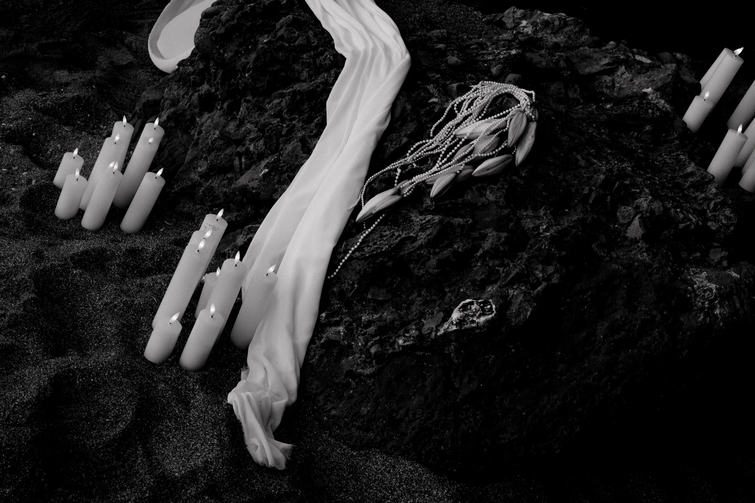 Bouquet de fleurs blanches, bougies allumées, et un tissu blanc posé sur une roche noire, en environnements sableux.