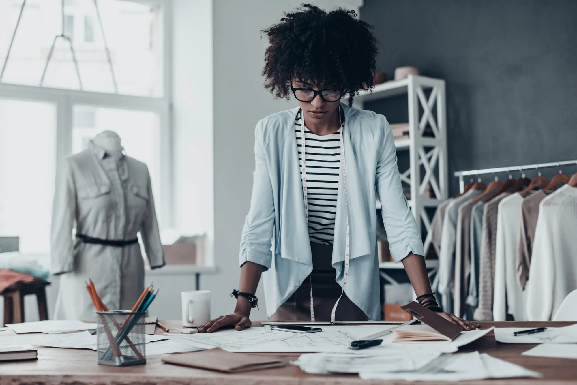 A woman in glasses and a striped shirt works at a large table with fabric and design sketches in a fashion design studio. The Little HR Department supports creative businesses and helps them to focus on being creative while we deal with the HR