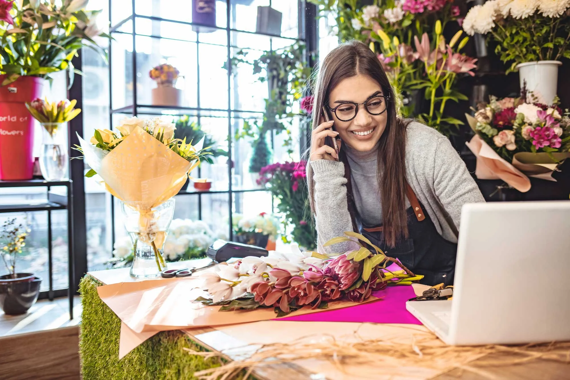 A young woman with glasses and long brown hair smiling while talking on her cellphone inside a flower shop. The Little HR Department can deliver great HR services to all types of businesses, including retail