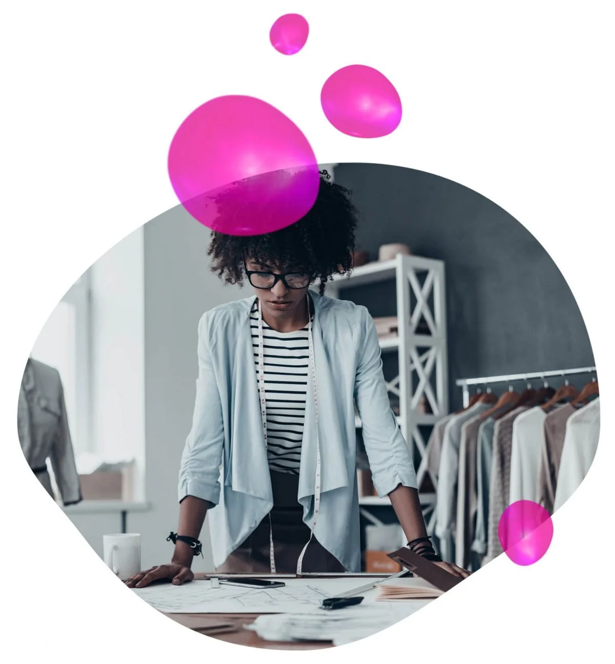 A woman with curly hair and glasses is standing at a table in a clothing design studio, reviewing sketches and fabric swatches, with clothing racks and shelves in the background.