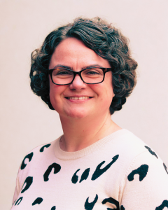 A woman with short, curly gray hair wearing glasses and a white sweater with black animal print, smiling at the camera. Her name is Michelle Potthurst