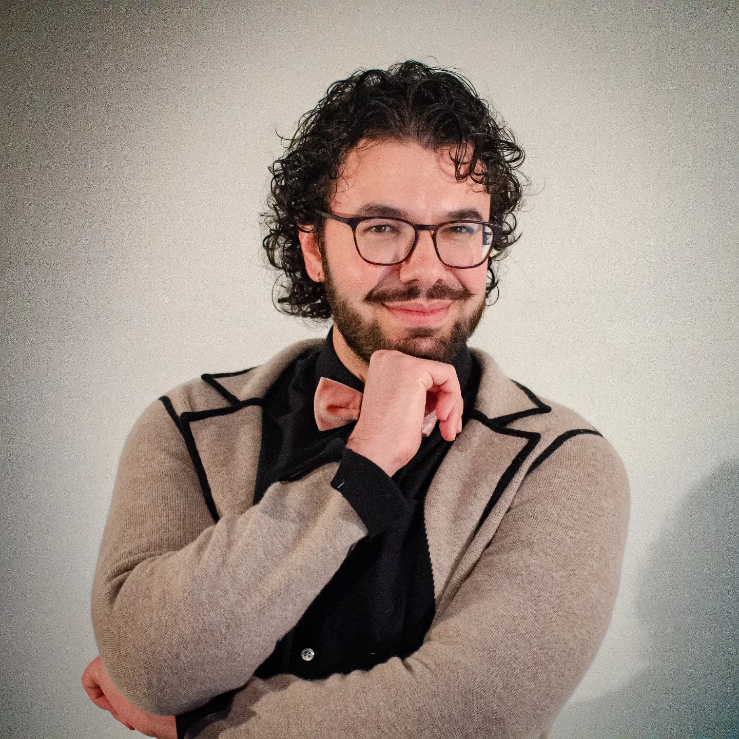 Jeune homme souriant, portant des lunettes, avec une moustache et une barbe, habillé d'un pull beige avec des lignes noires, une chemise noire et un nœud papillon rose, posant avec la main sous le menton contre un fond blanc.