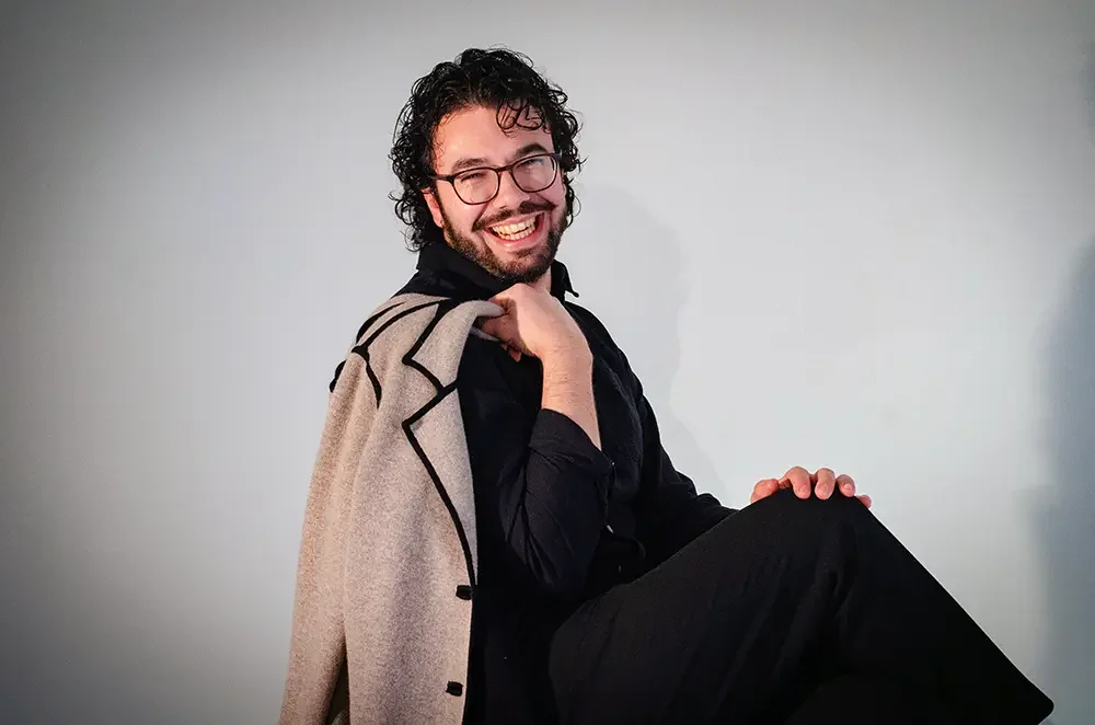 Homme souriant avec lunettes, portant une veste beige sur l'épaule, assis contre un mur blanc.