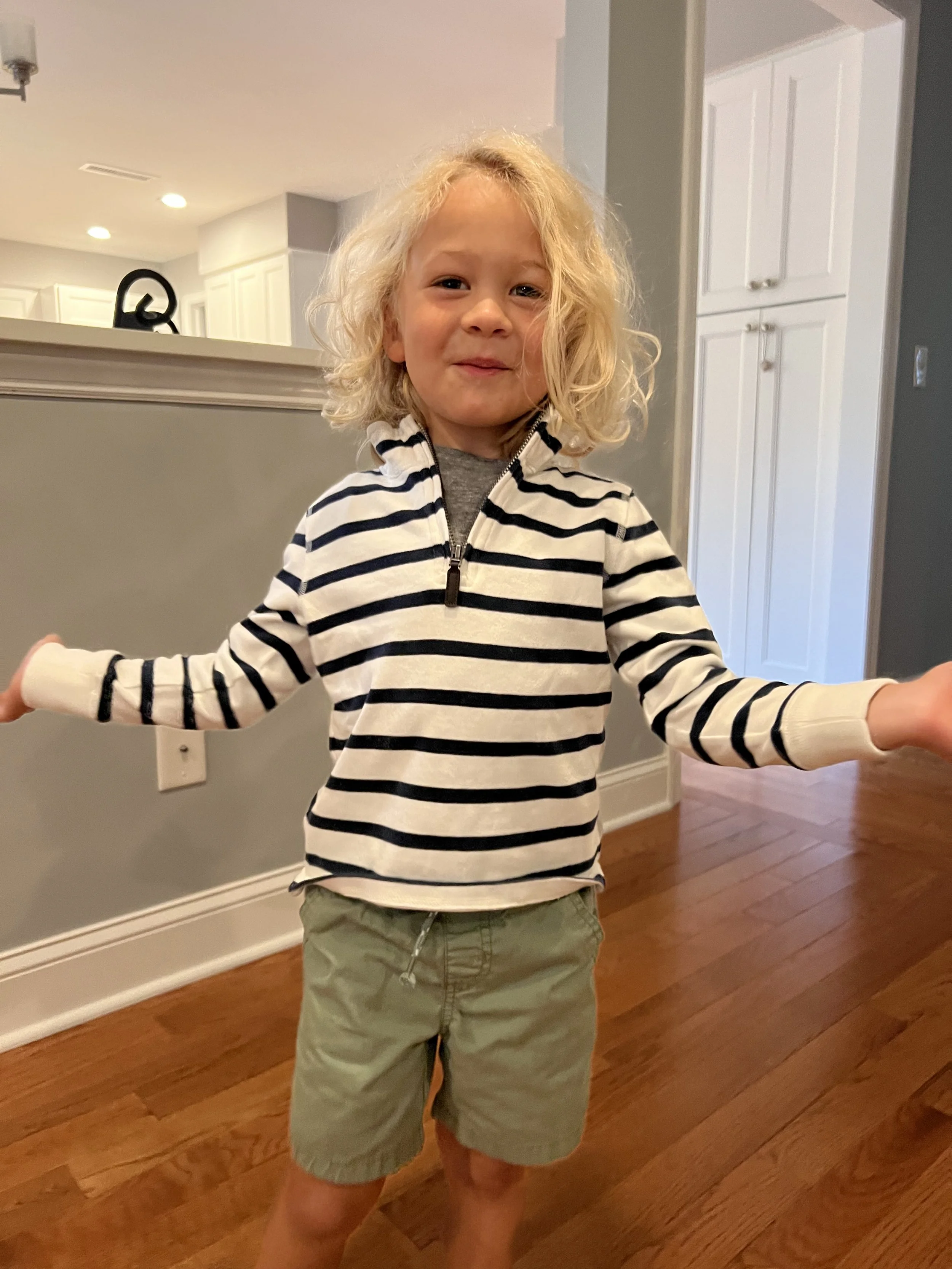 A young child with curly blonde hair wearing a striped jacket with arms outstretched standing in a house with wooden floors and white cabinets.