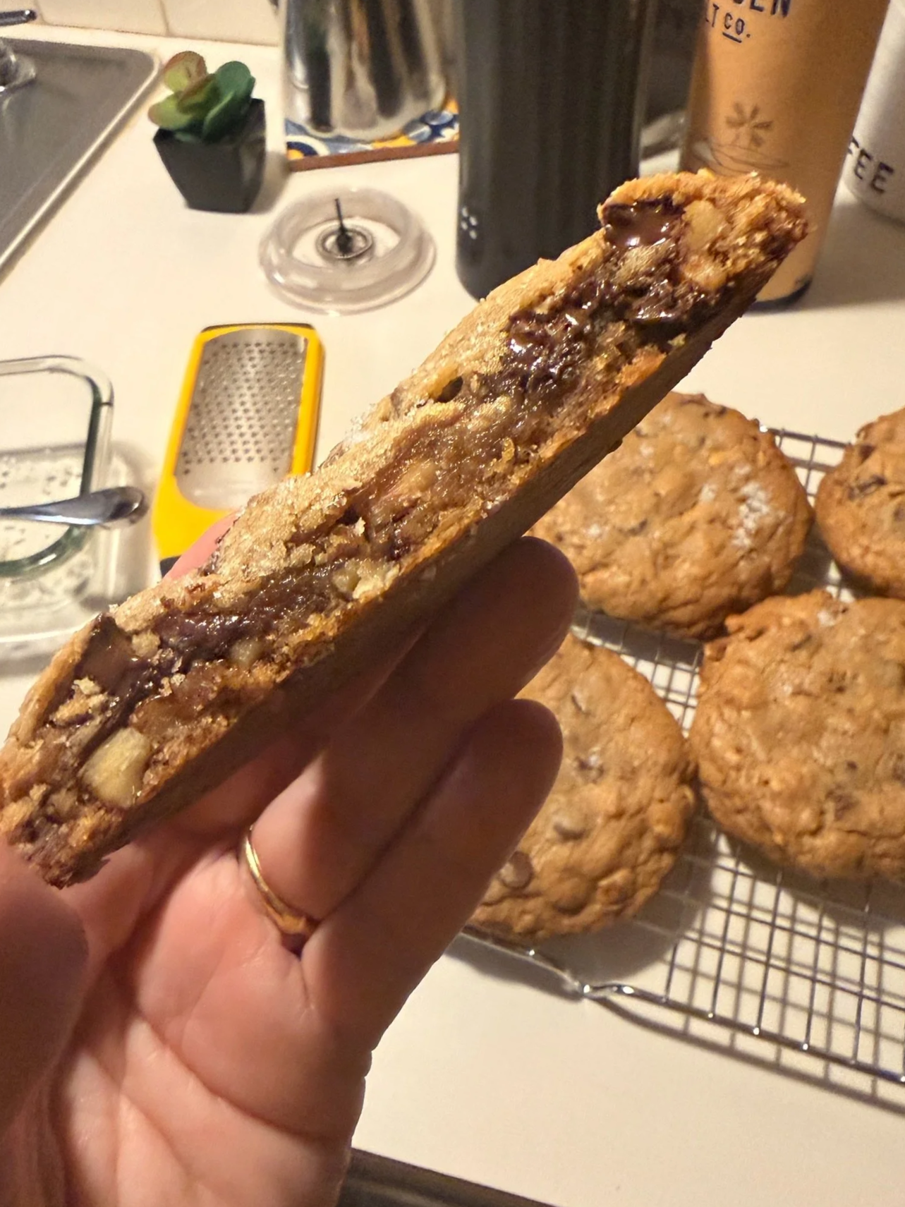 Close-up of a person's hand holding a cookie with chocolate and nuts, with more cookies cooling on a wire rack in the background on a kitchen countertop.