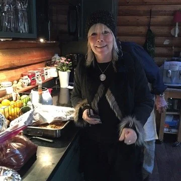 Woman in a black beanie and dark jacket in a cozy kitchen, smiling and holding a phone, with food and kitchenware on the counter.