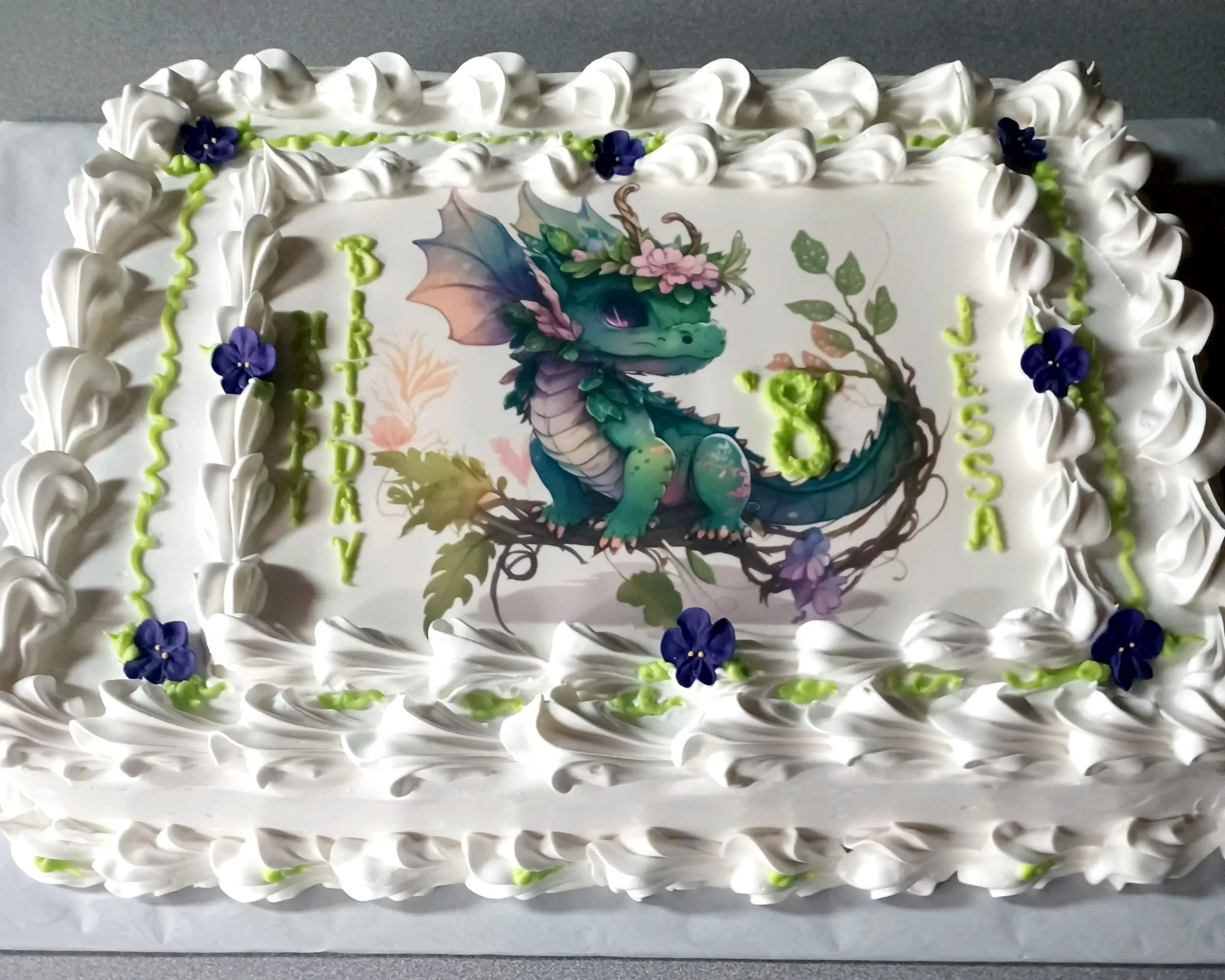 A rectangular birthday cake decorated with white icing, purple flowers, and green piping, featuring an edible image of a cartoon dragon with wings, horns, and a crown of pink flowers sitting on a branch.