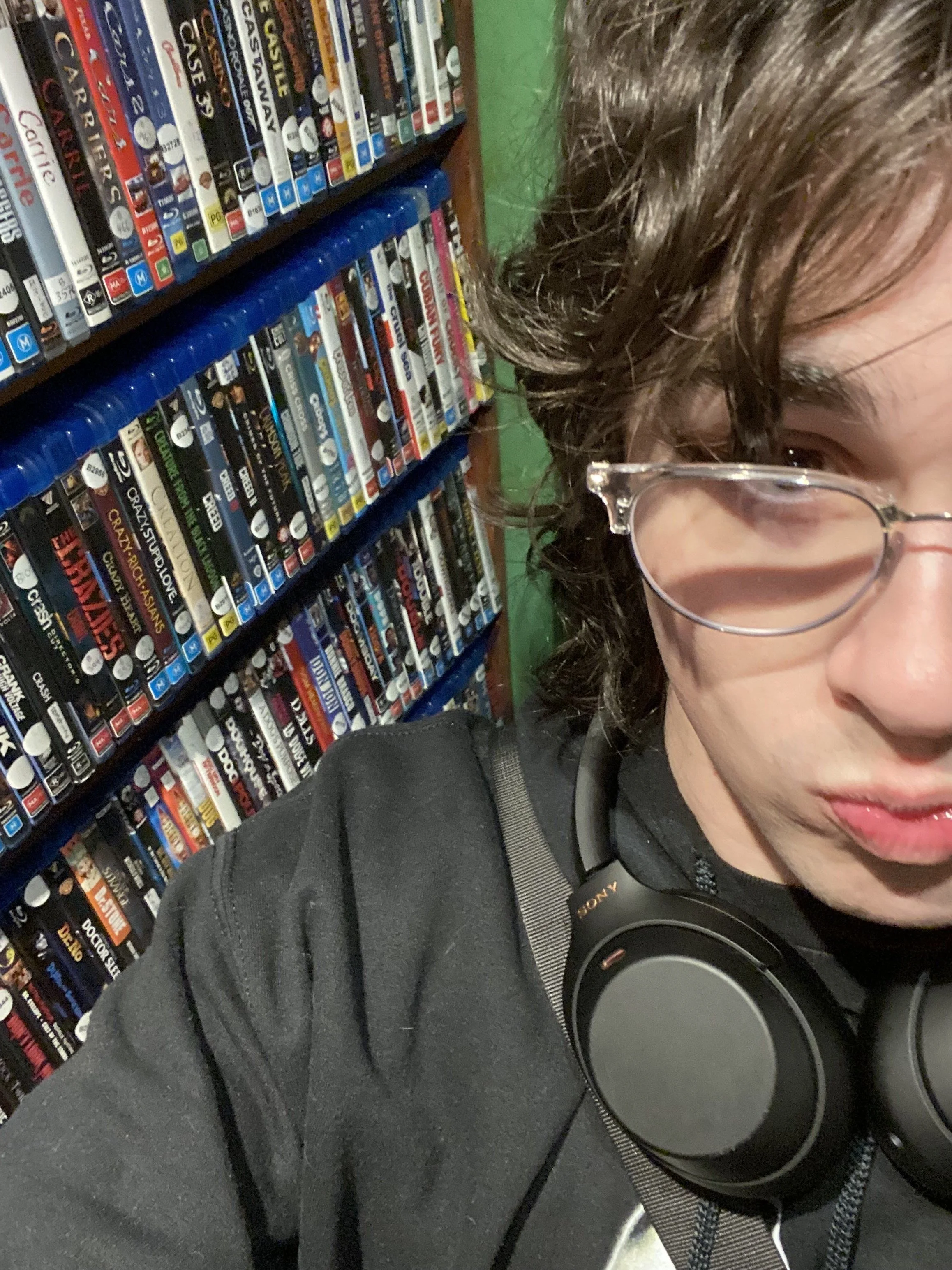 A person with curly hair wearing glasses and headphones, standing in front of shelves filled with DVDs.