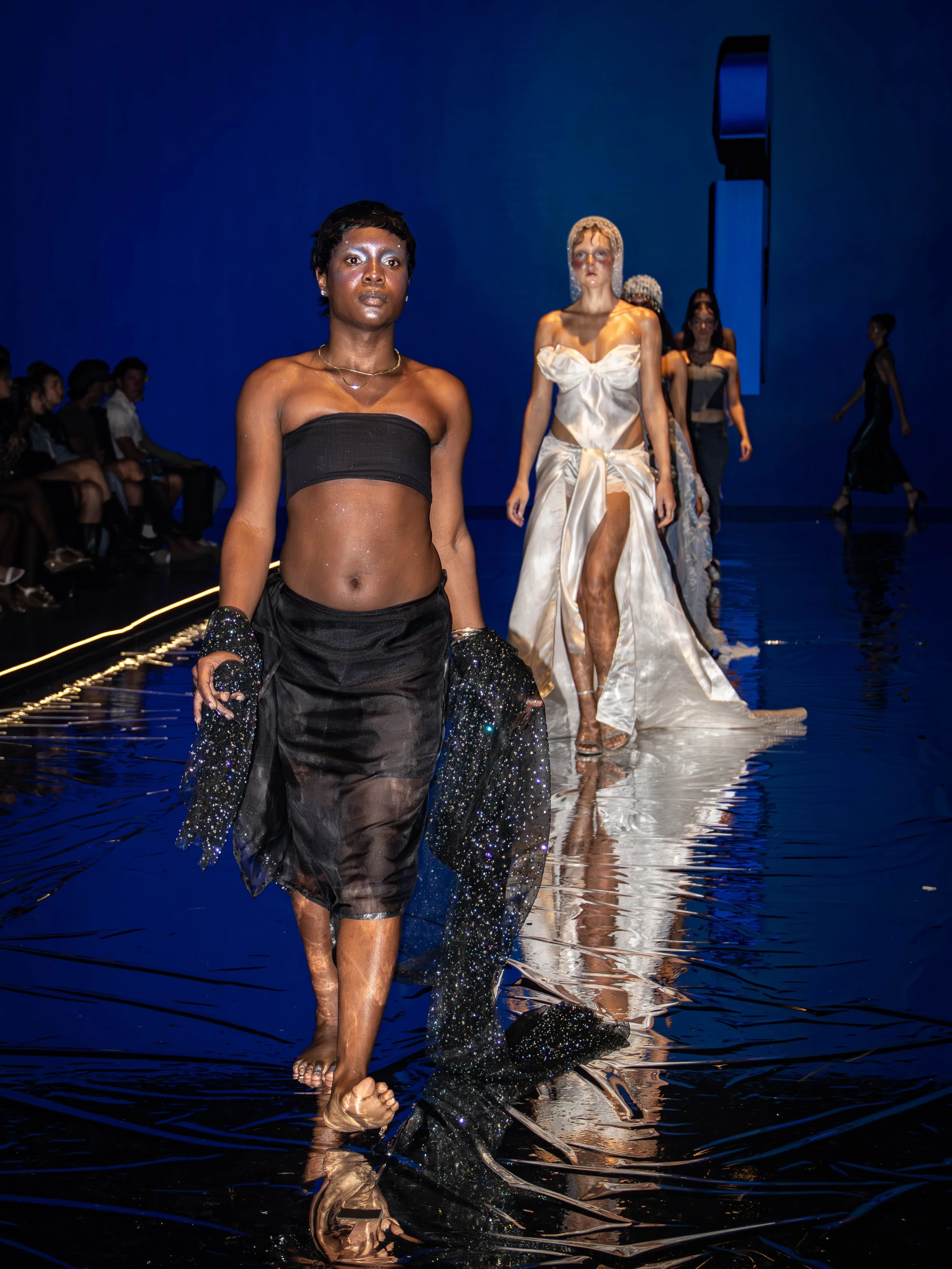 Models walking down a runway waterwalk at a fashion show, with audience seated on side and a blue background.