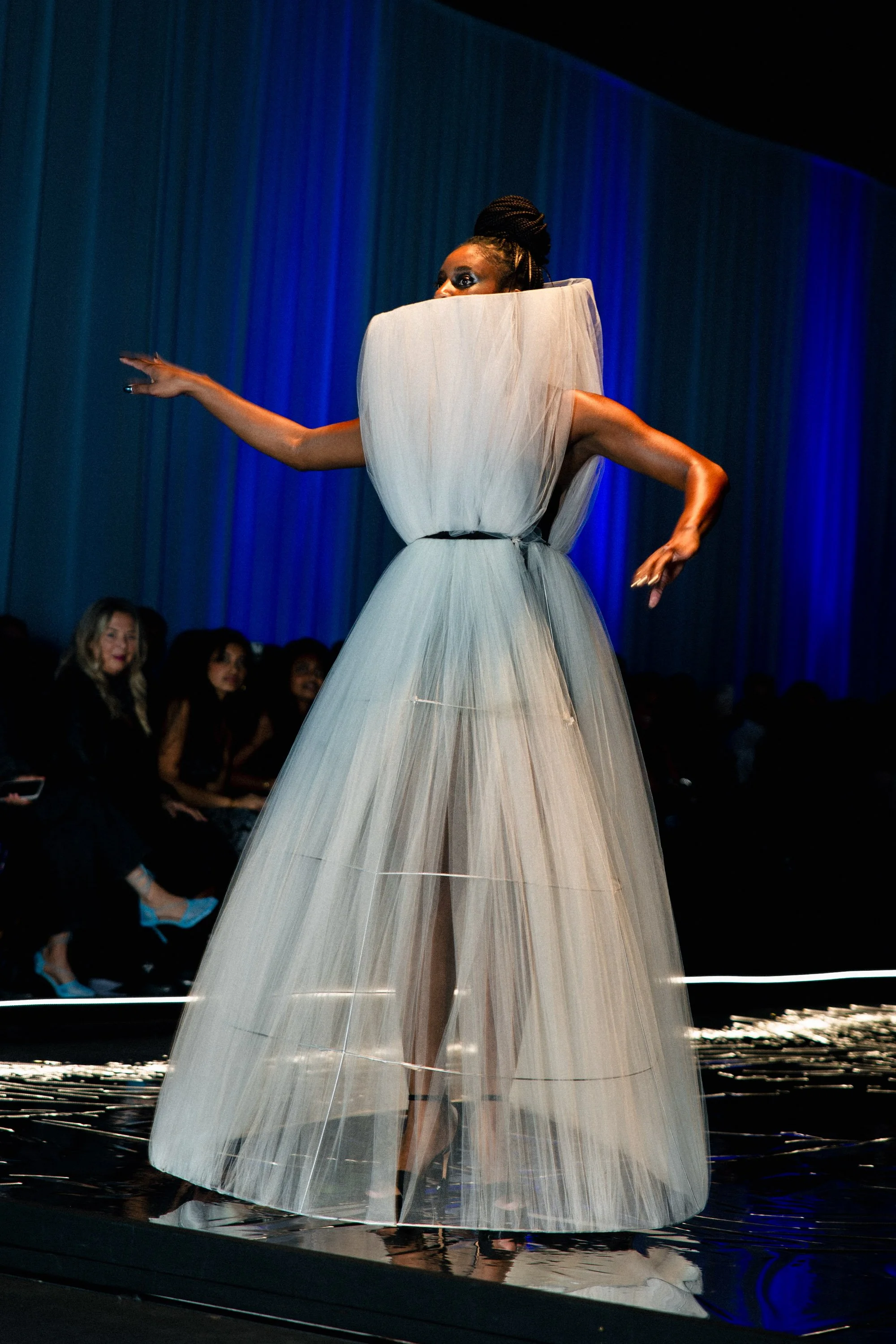 Model walking on runway wearing a flowing, voluminous gown with sculptural shoulder detail, audience seated beside runway.