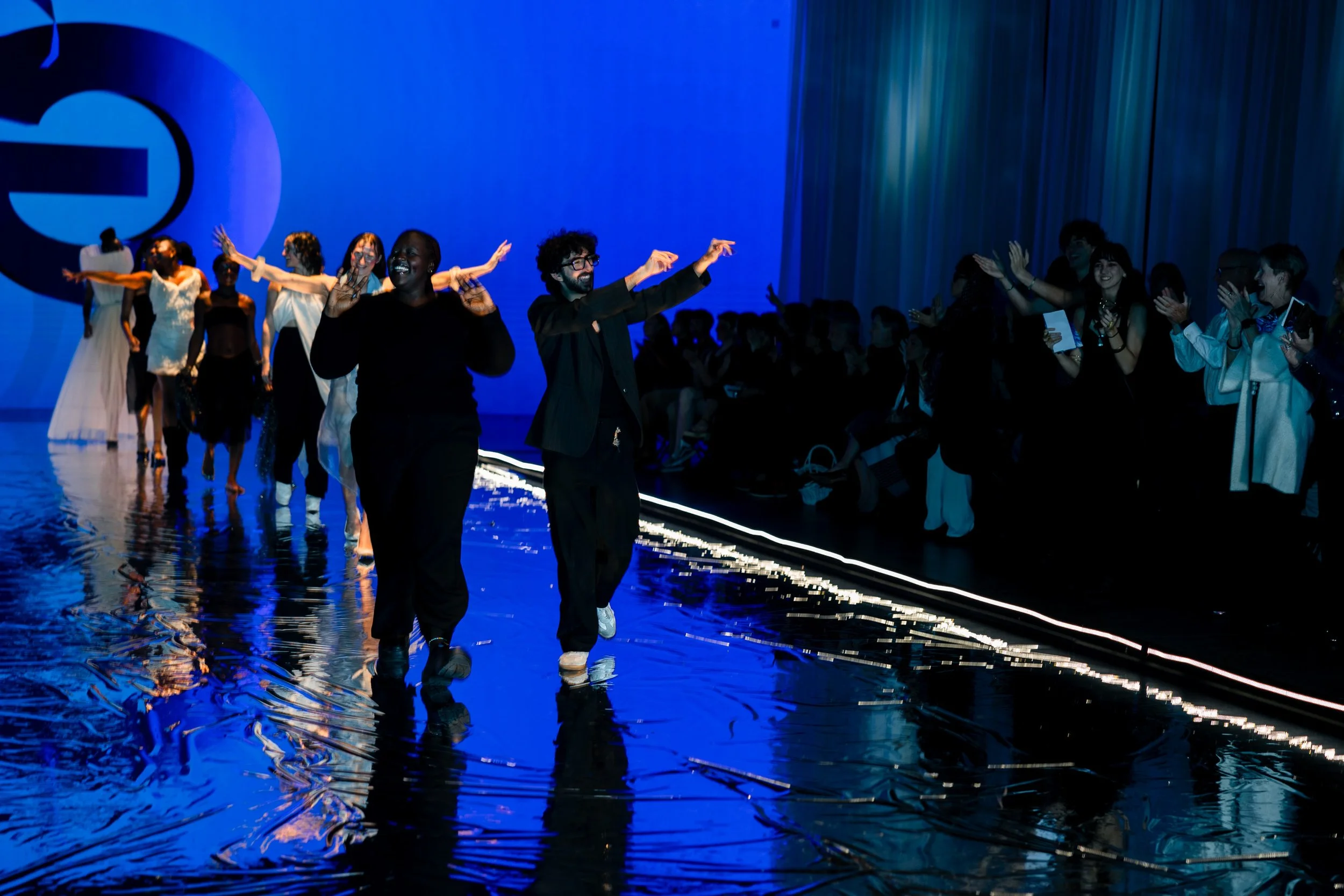 Fashion show runway with models walking and audience clapping, illuminated by blue and purple lighting.