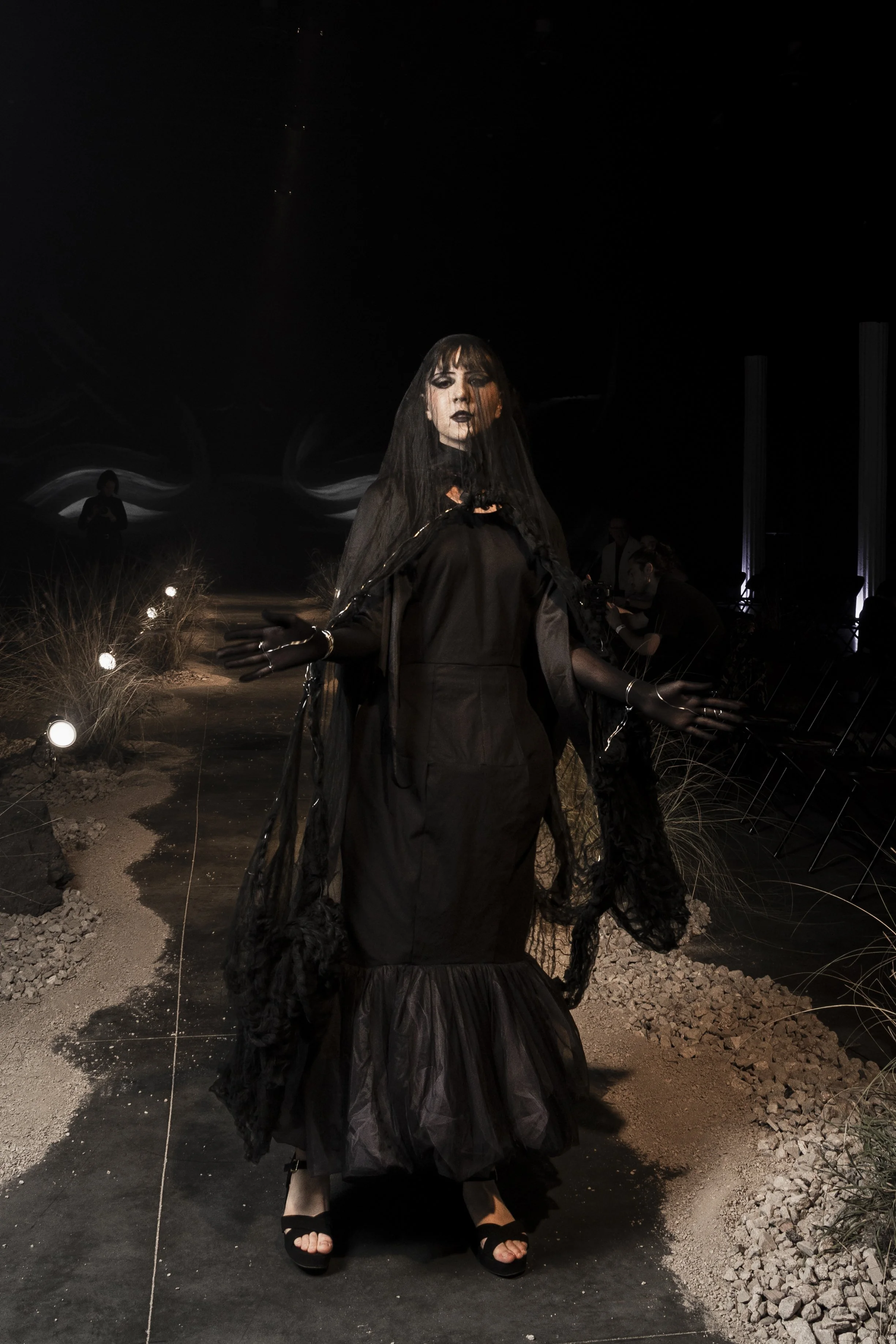 A woman dressed in black gothic attire with dark makeup and a veil stands in a dark, indoor setting with minimal lighting and desert-themed decorations.