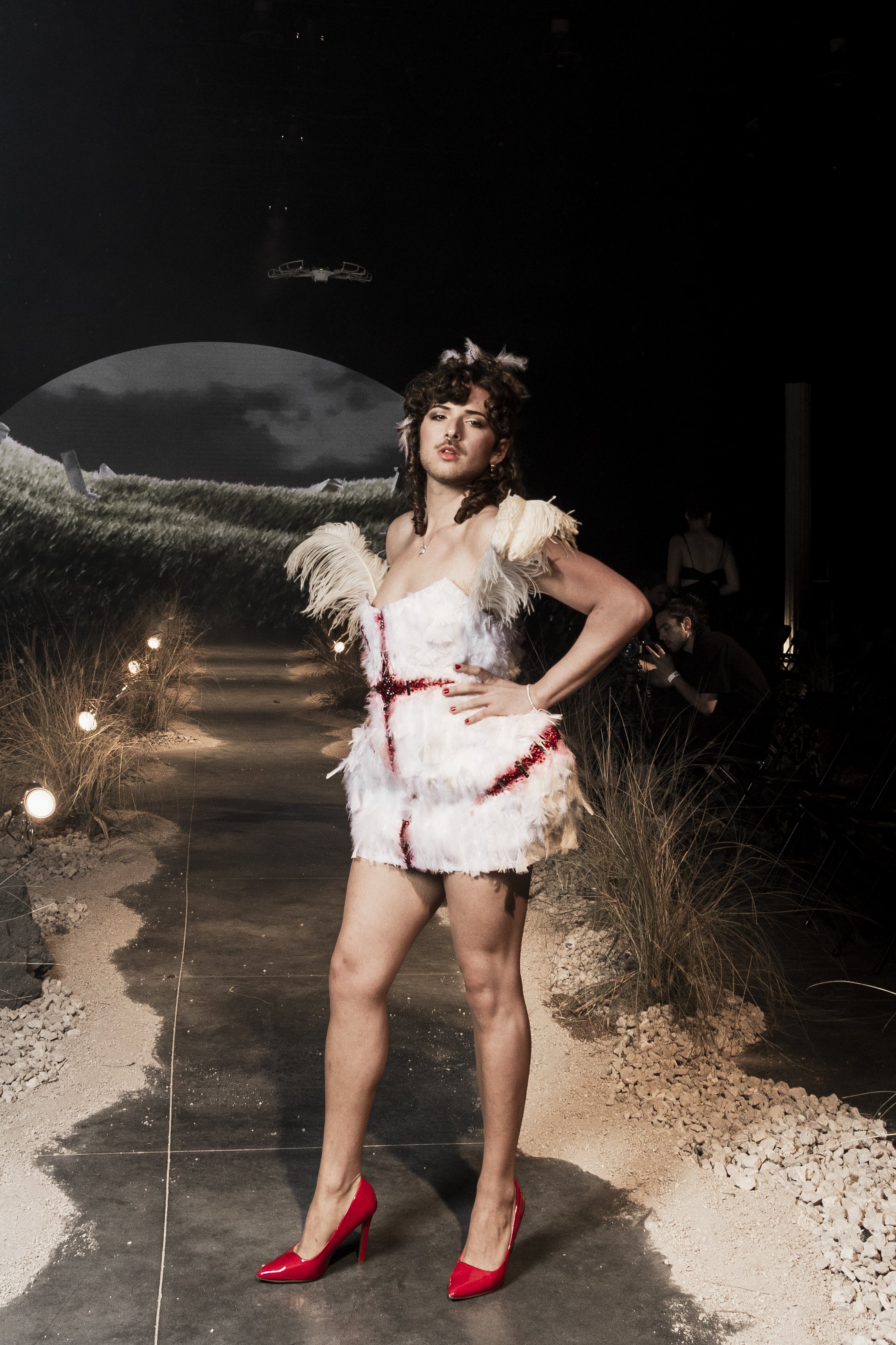 A woman with curly hair standing on a runway, wearing a white feathered dress with red accents, red high heels, and posing with her hand on her hip. The background depicts a night sky with a spaceship and a large moon, simulating a lunar landscape wi