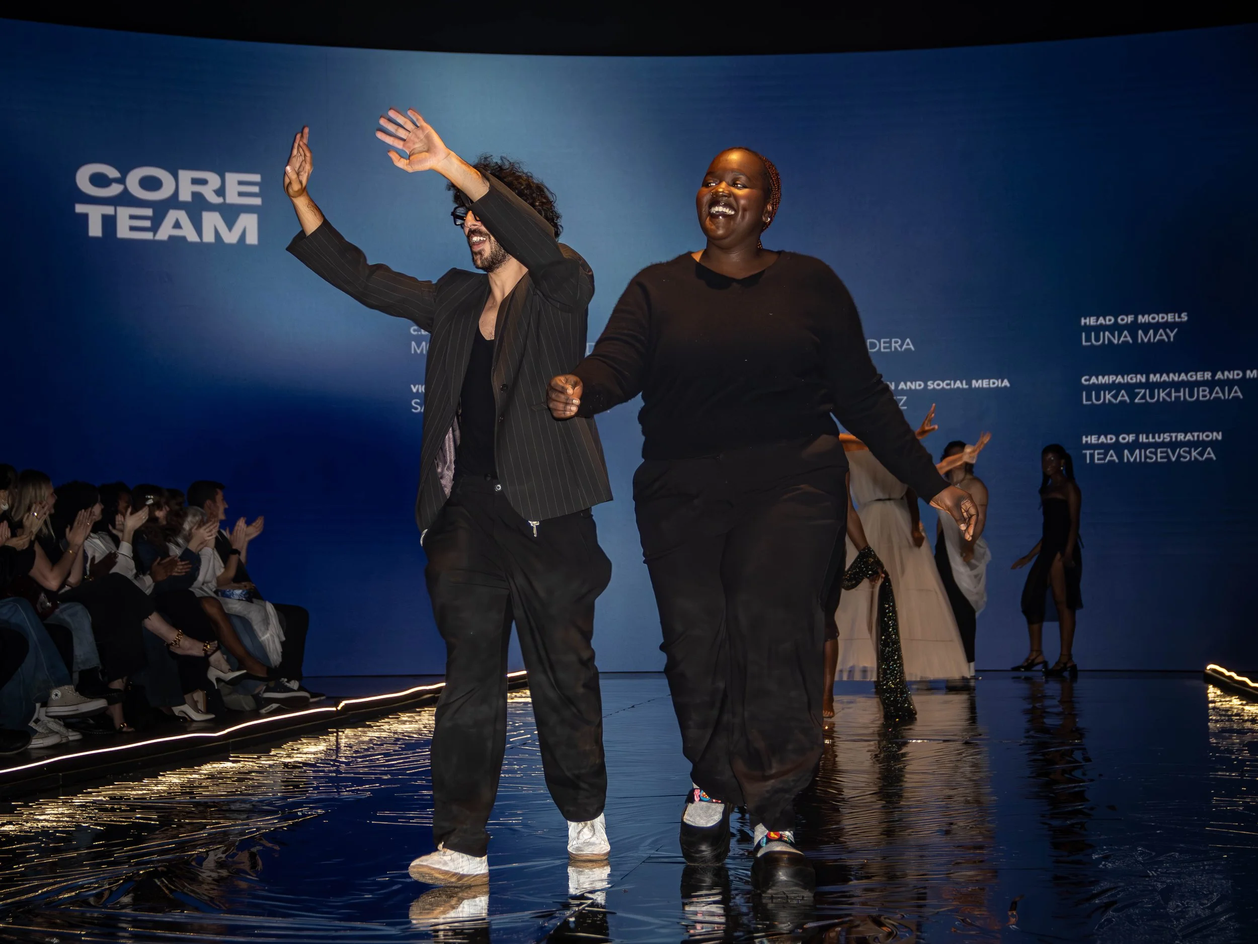 Two fashion models walking down the runway during a fashion show, with a blue backdrop that reads 'CORE TEAM', surrounded by seated audience members and other models in the background.
