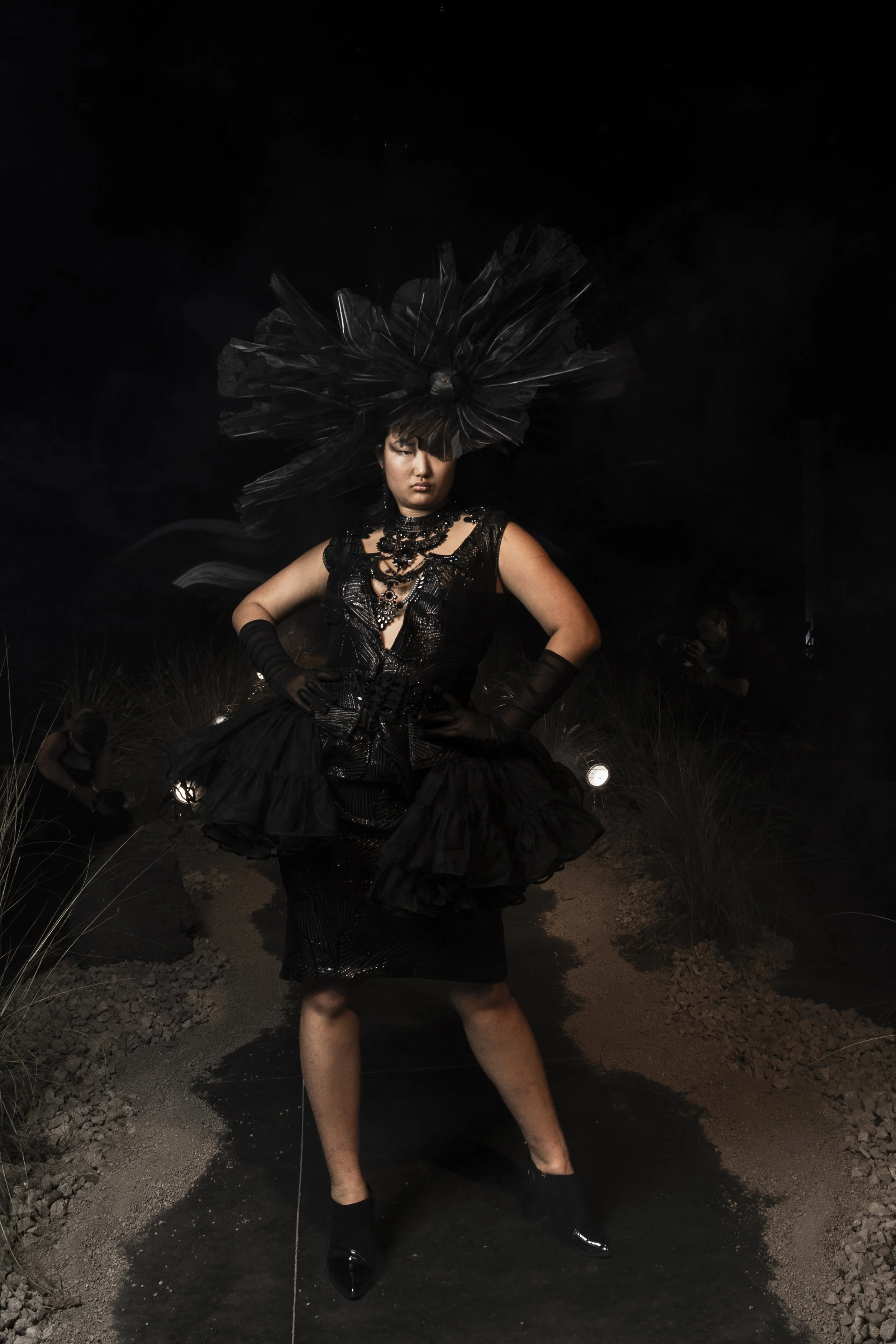 A woman dressed in an elaborate black gown with intricate jewelry and a large black headpiece, standing outdoors at night with her hands on her hips, with other people in the background.