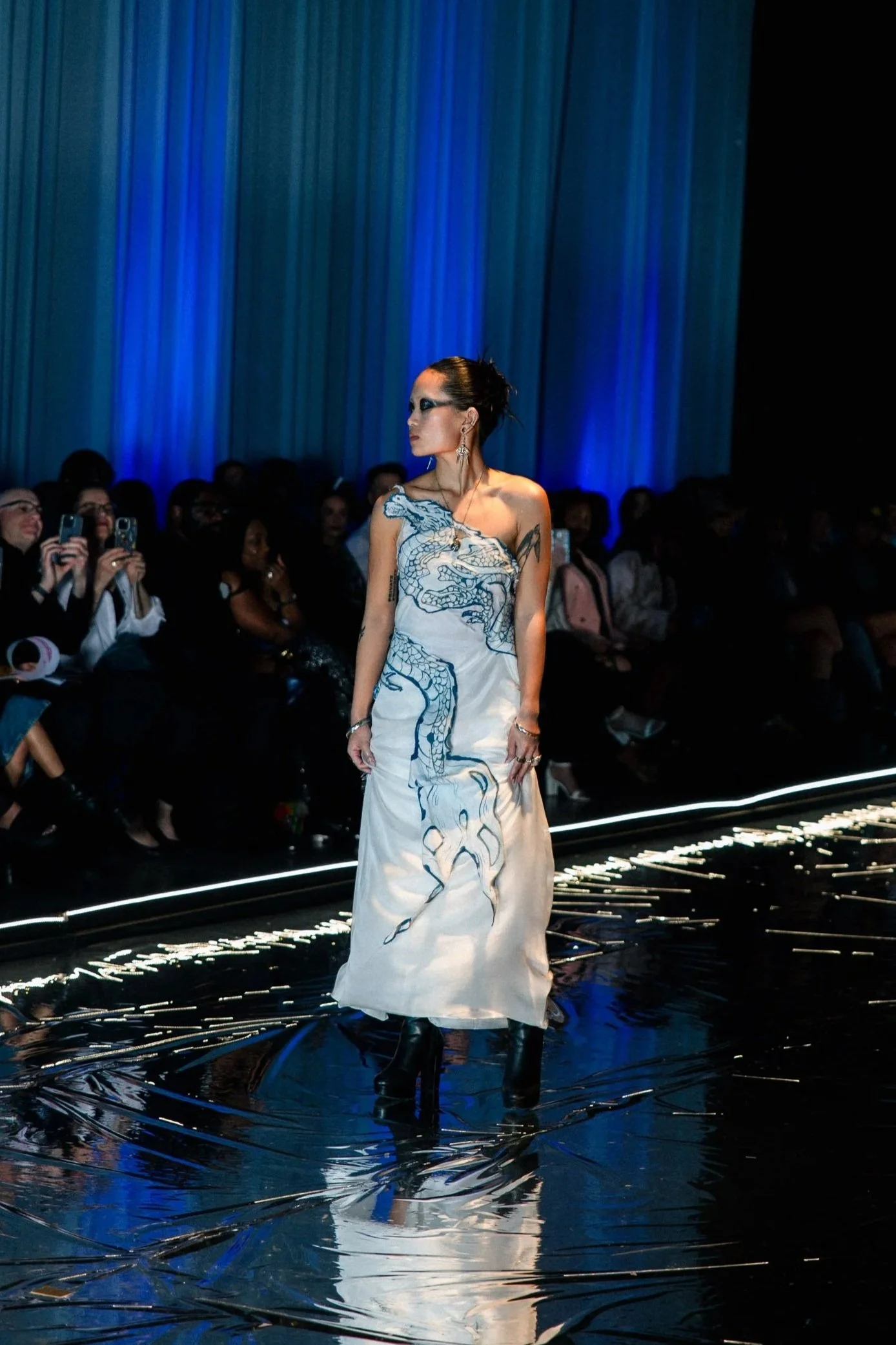 A model walks on a reflective runway during a fashion show, wearing a white dress with a blue dragon design, black high-heeled boots, and bold makeup, with an audience watching in the background.