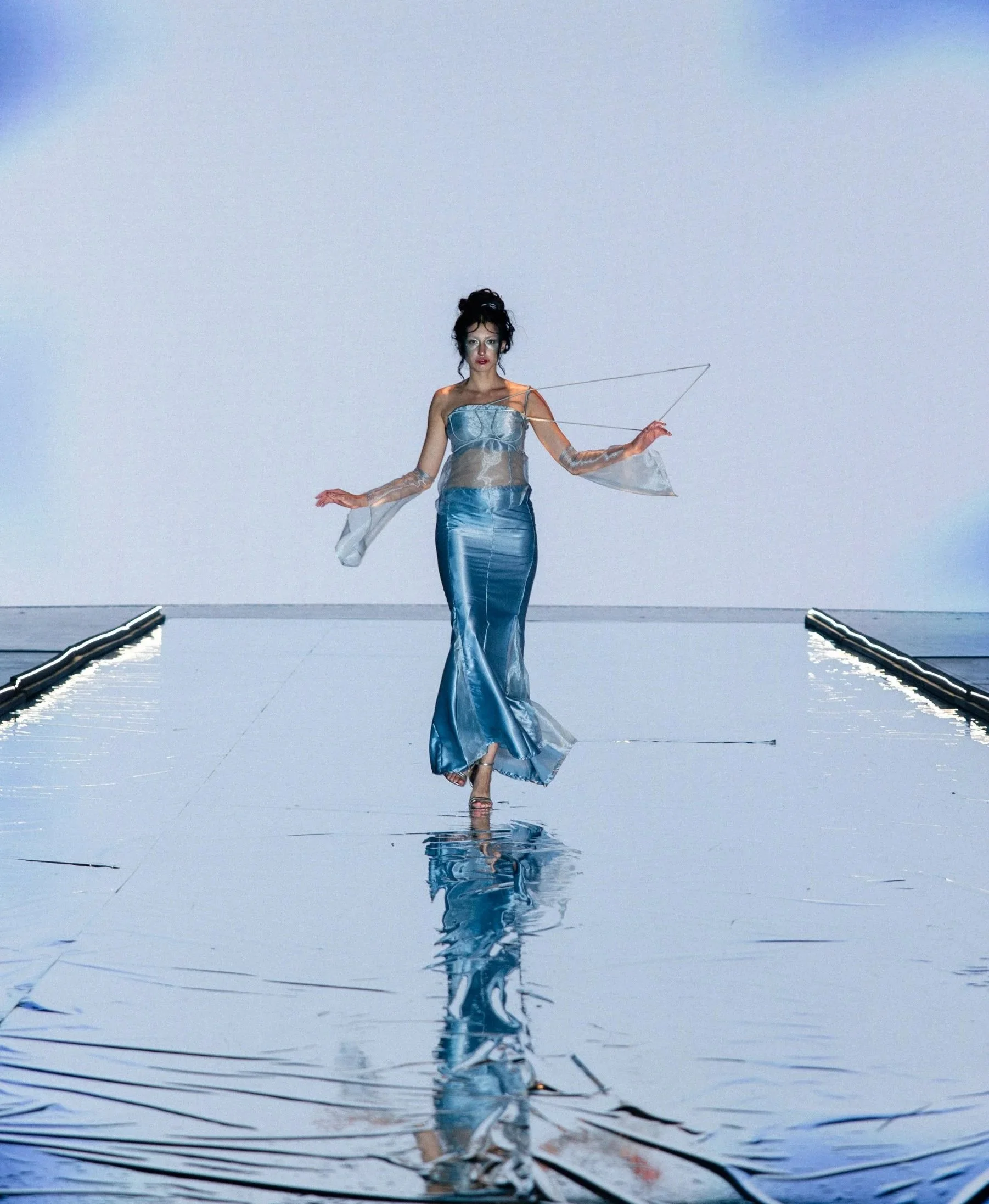 A woman walking on a reflective runway over water, wearing a blue satin gown and transparent sleeves, with a cloudy sky in the background.