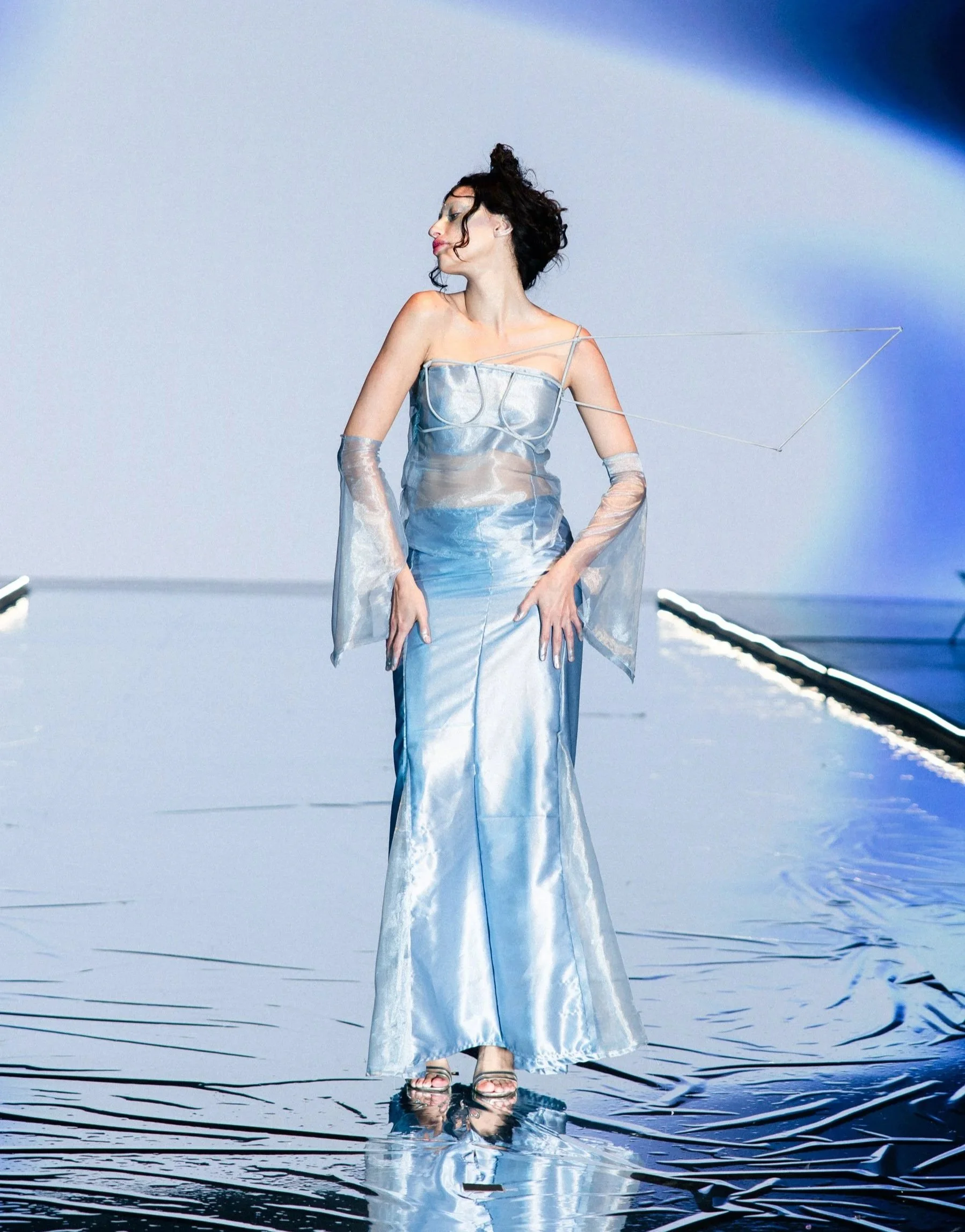 A woman in a shiny silver dress and transparent gloves walks on a reflective, metallic runway with a blue and white background.