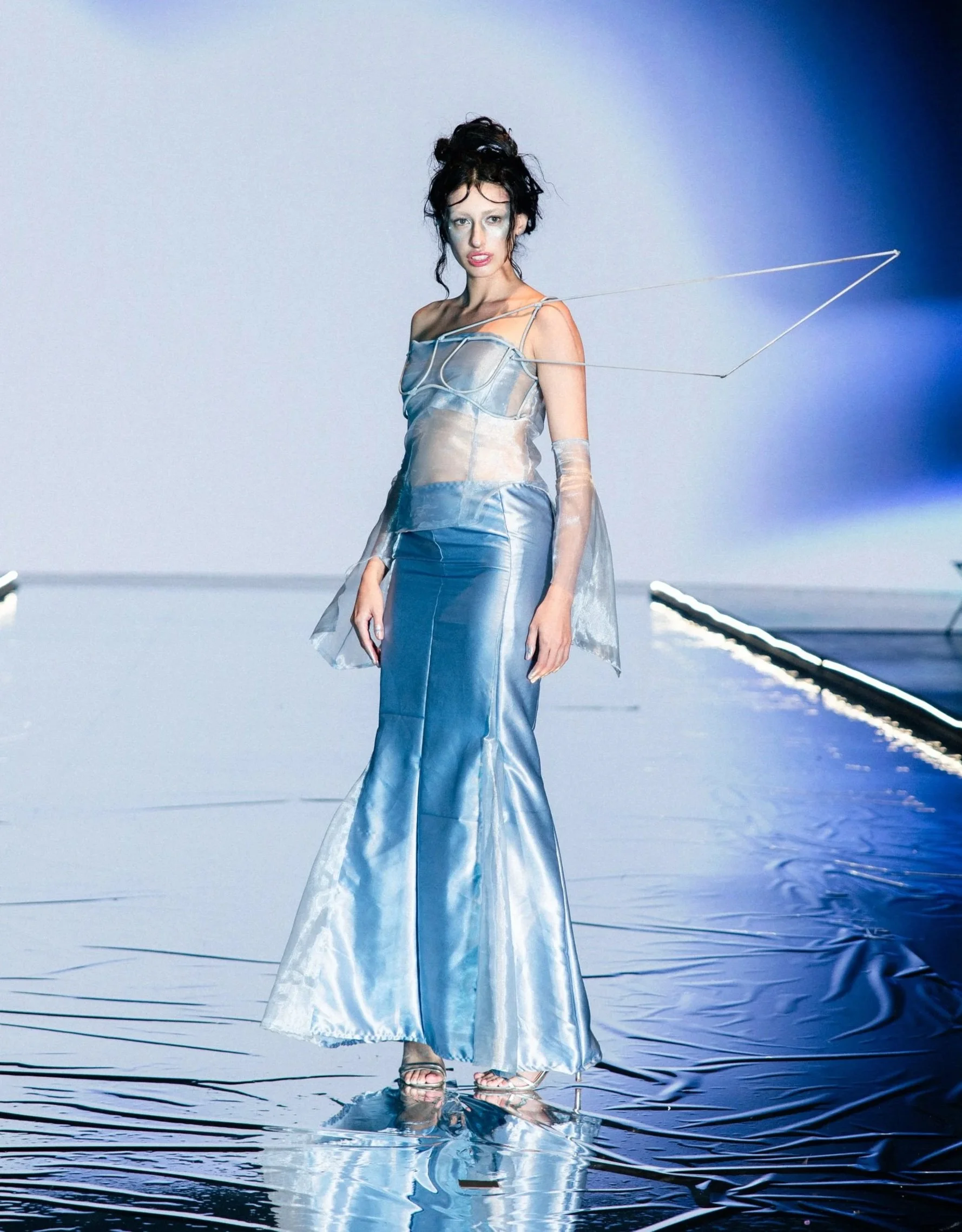 A woman in a shiny, iridescent gown with a fitted bodice and long, flowing skirt standing on a reflective surface with a blue and gray background.