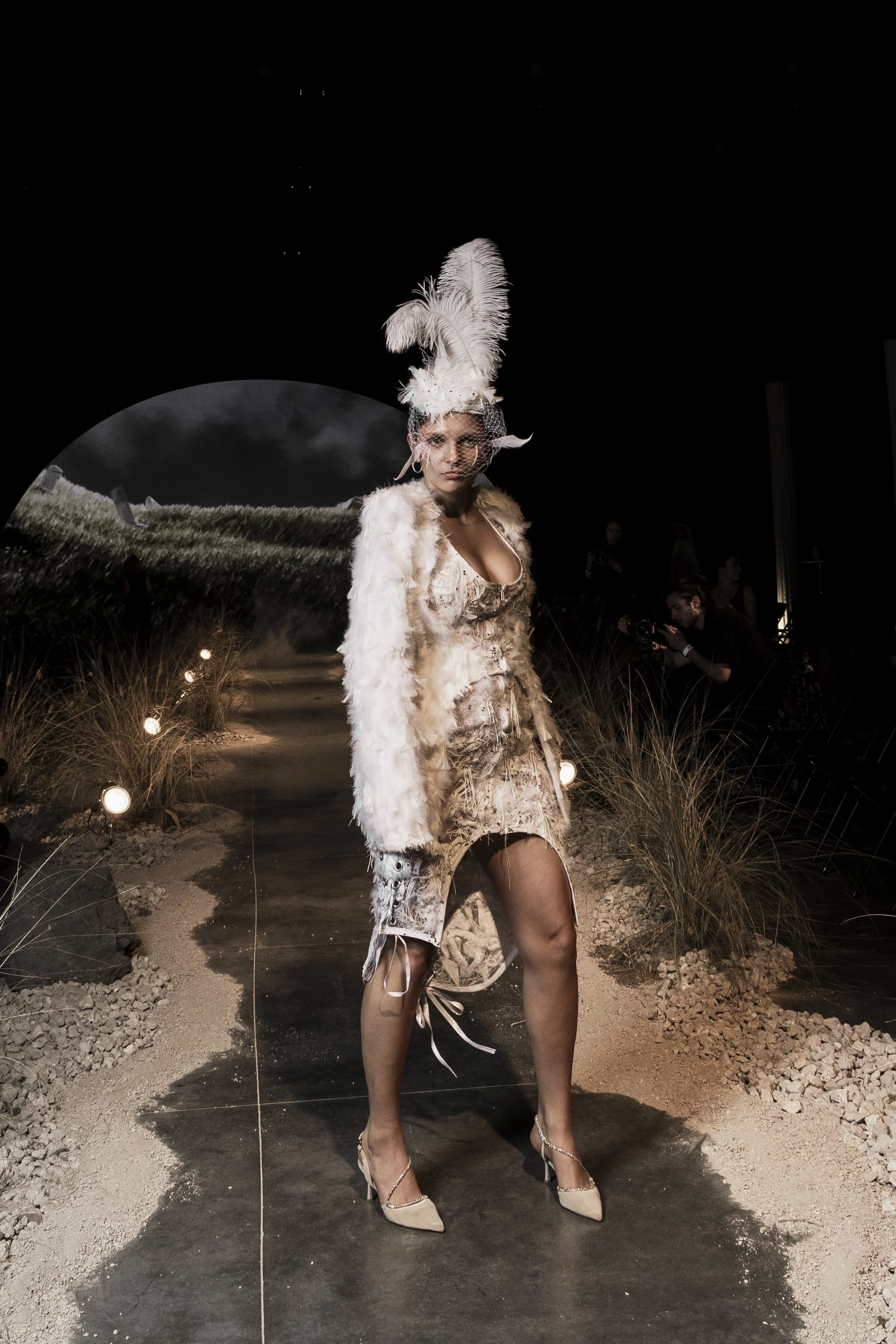 A fashion model walking on a runway, wearing a feathered headpiece, a fur-like coat, and high heels, with a desert landscape backdrop at night.
