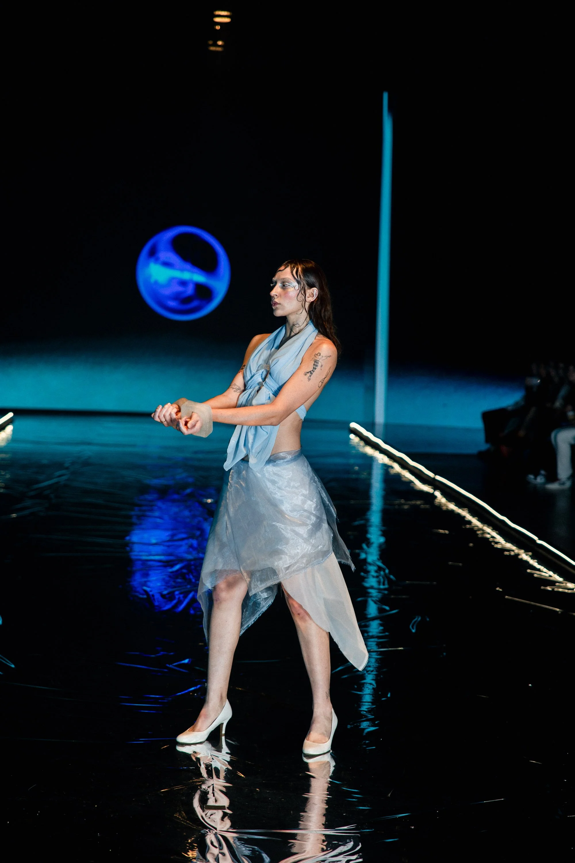 Model walking on a runway during a fashion show with a dark backdrop, blue abstract globe, and illuminated runway edges.