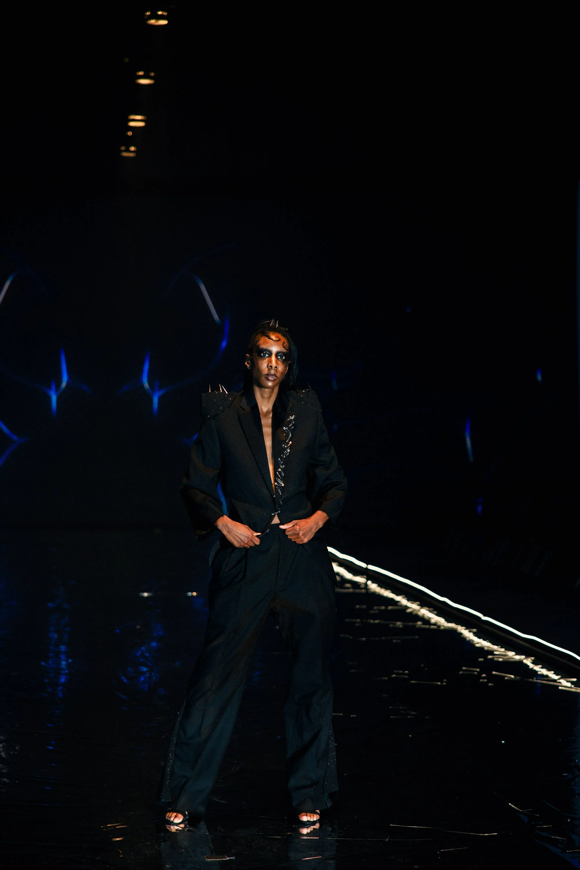 Fashion model walking on runway wearing black blazer and wide-legged pants with spikes on shoulders, in a dark setting with neon lights reflecting on the floor.