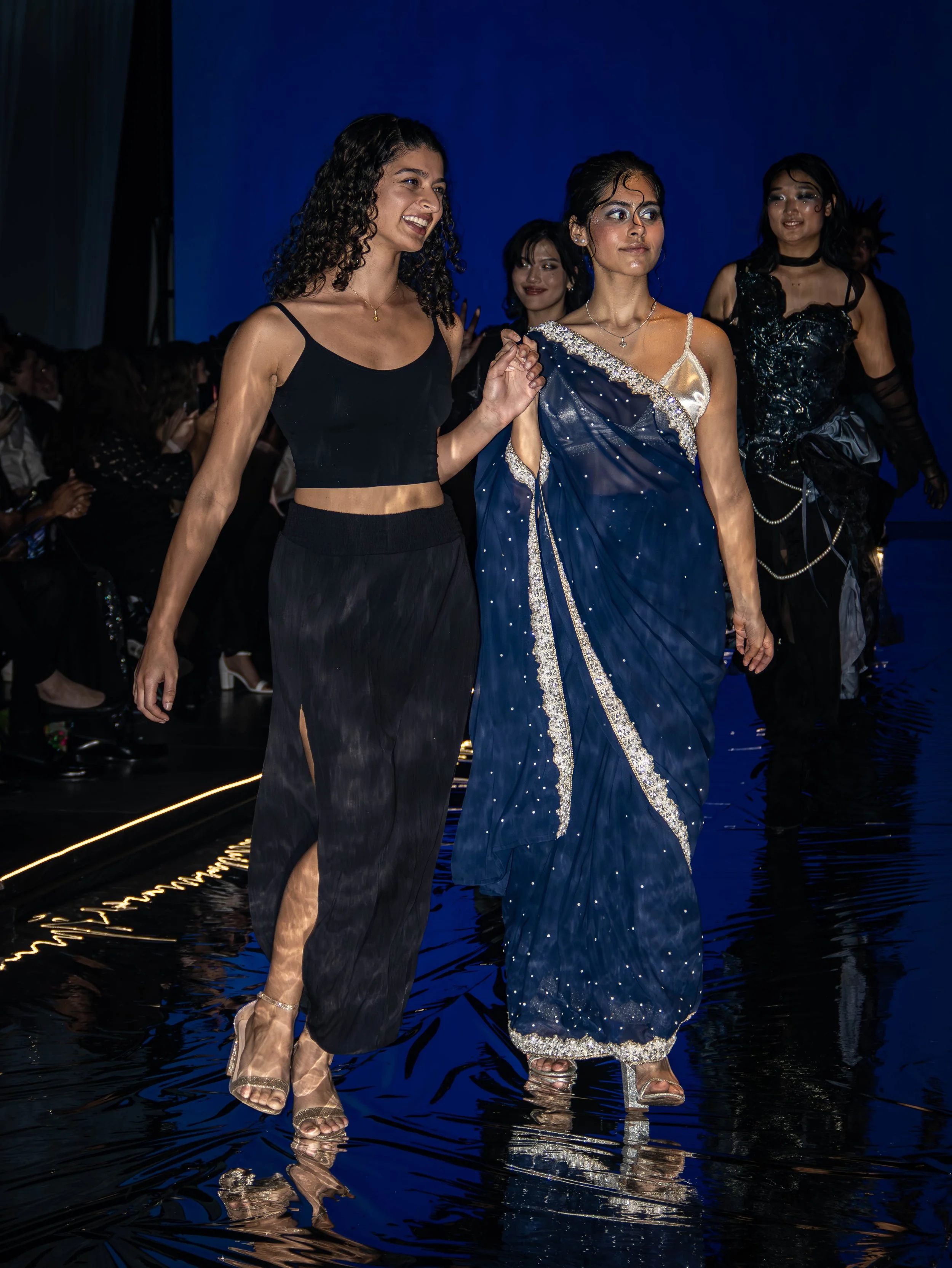 Two women walk on a runway during a fashion show, with bright lighting and a dark background. The woman on the left is wearing a black crop top and loose black pants with a slit, and the woman on the right is wearing a blue sari with silver embellish