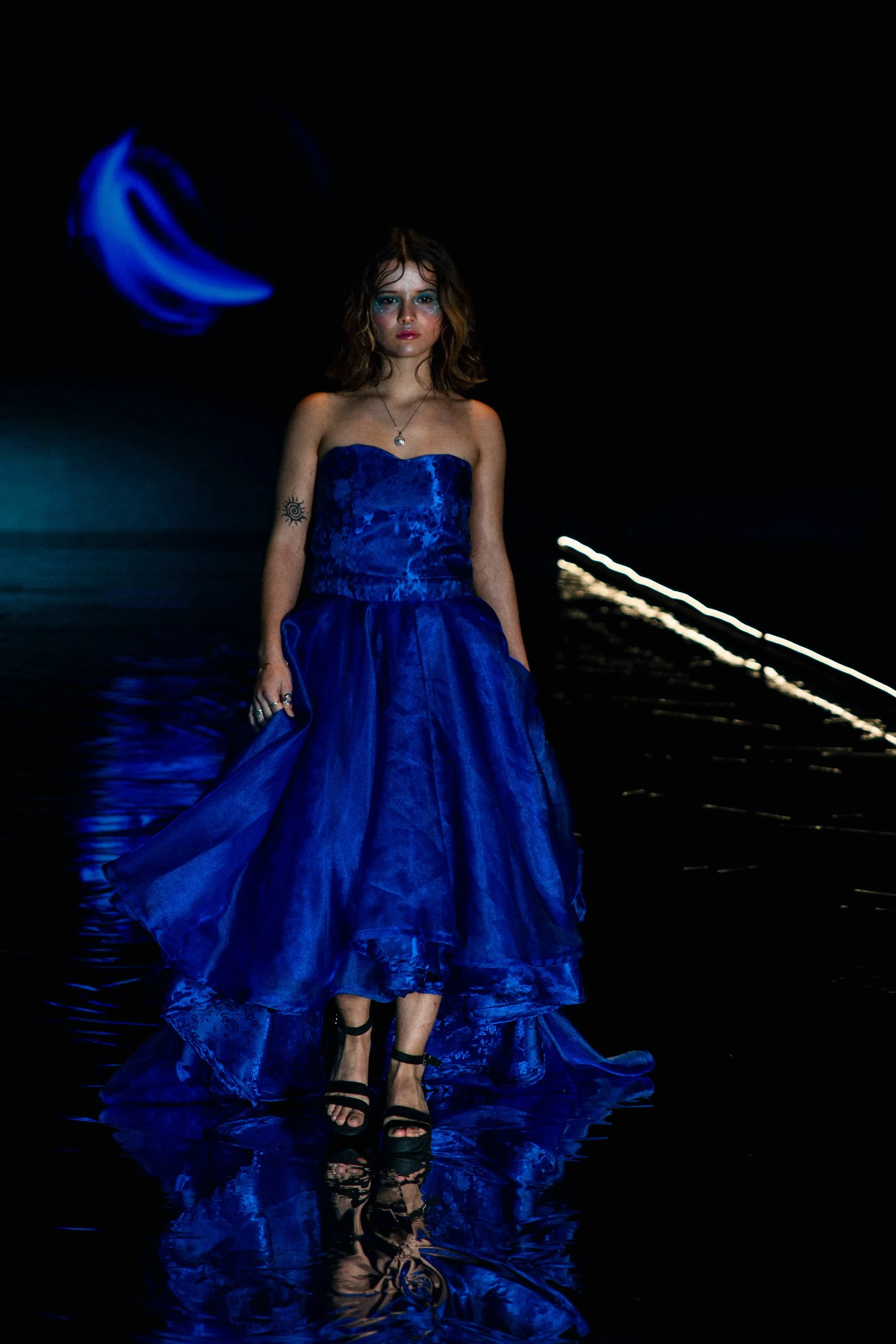 A woman walking on a runway reflecting water, wearing a strapless blue gown and black high heels, with makeup and hair styled in loose waves, dark background, and blue and white lighting effects.