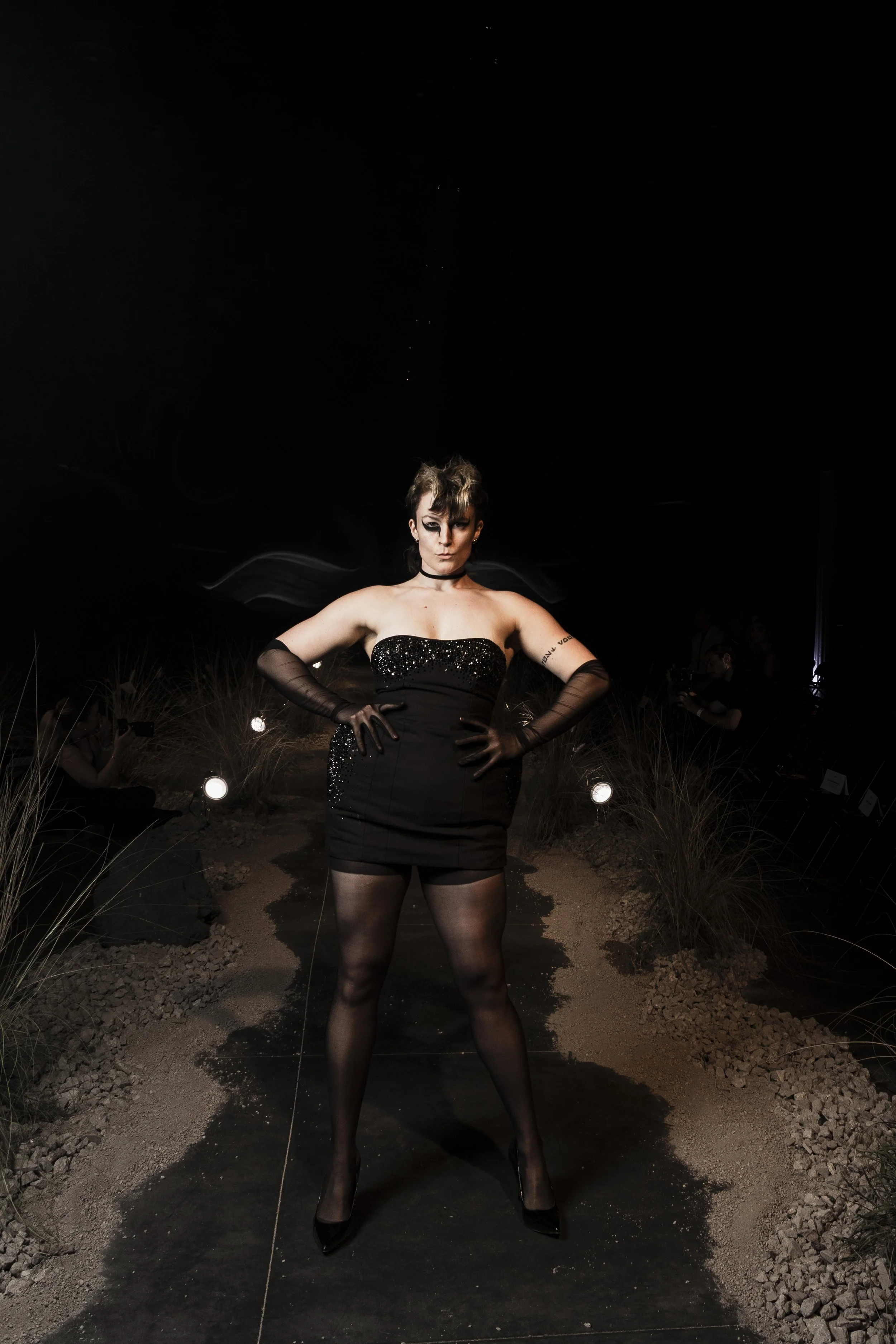 A woman in a black, sparkly strapless dress and black sheer gloves stands confidently with her hands on her hips on a dark, illuminated runway with dry grass and rocks, and a starry night sky overhead.