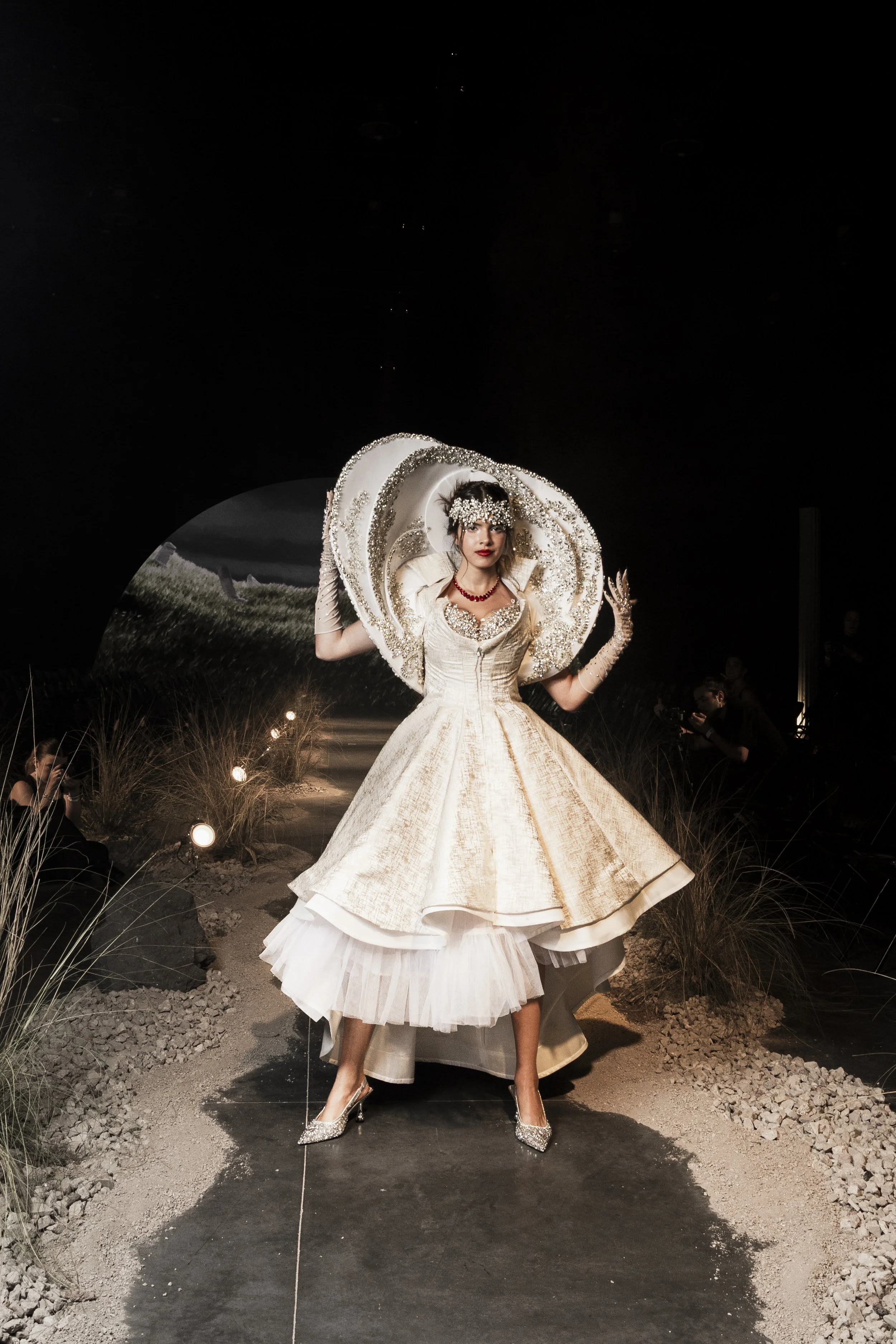 A woman in an elaborate white wedding gown with a wide skirt and lace details, wearing high heels, stands on a runway with a dark background. She has a large, decorated headpiece and jewelry, and there are photographers and onlookers capturing the mo